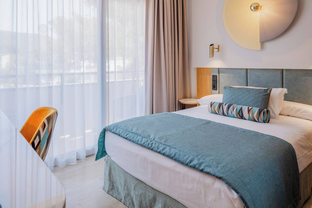 Una habitación de hotel con cama, una silla, una mesita de noche y una lámpara de pared.