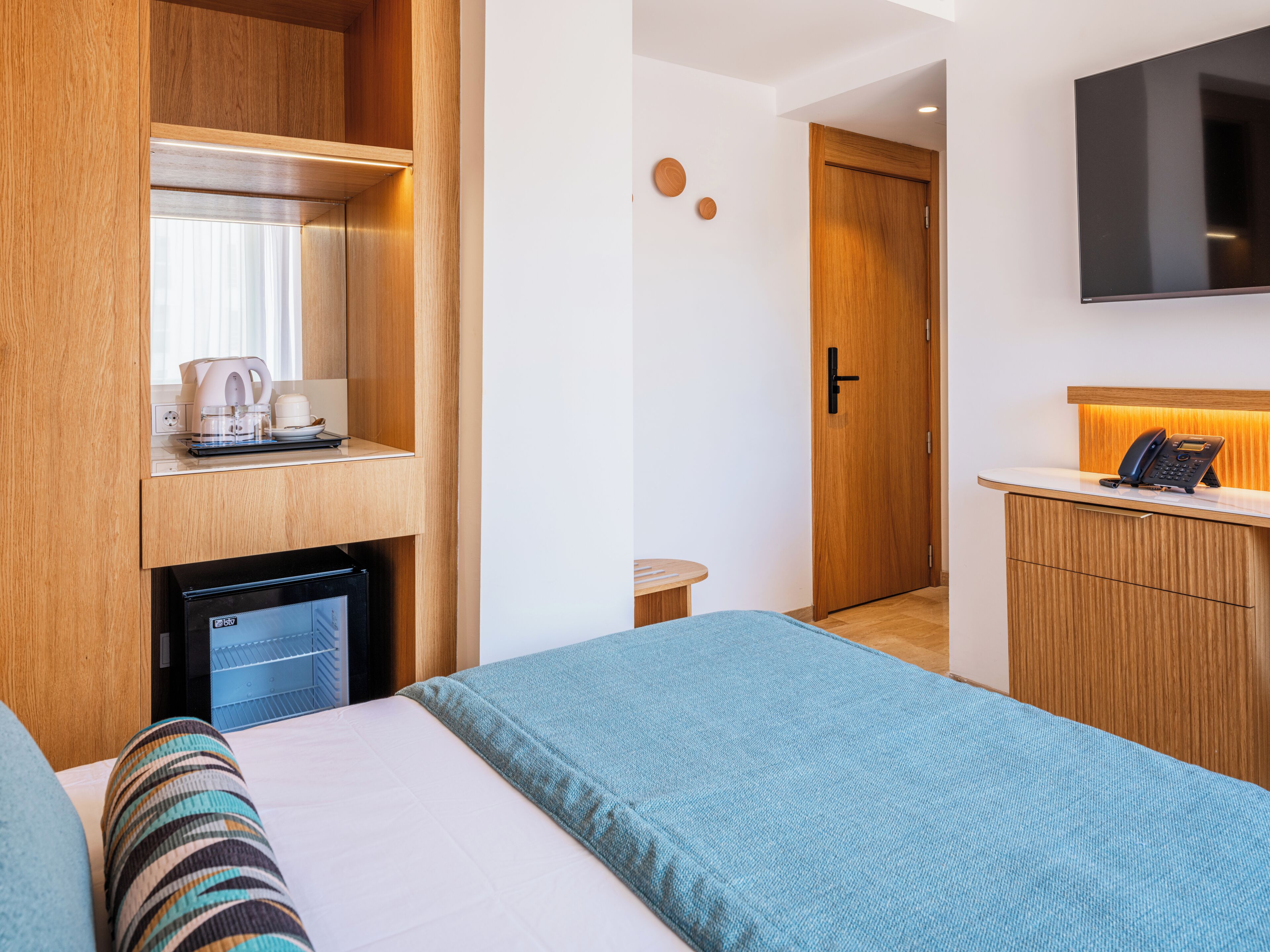 Habitación de hotel con cama, escritorio con teléfono, refrigerador pequeño y televisor montado en la pared.