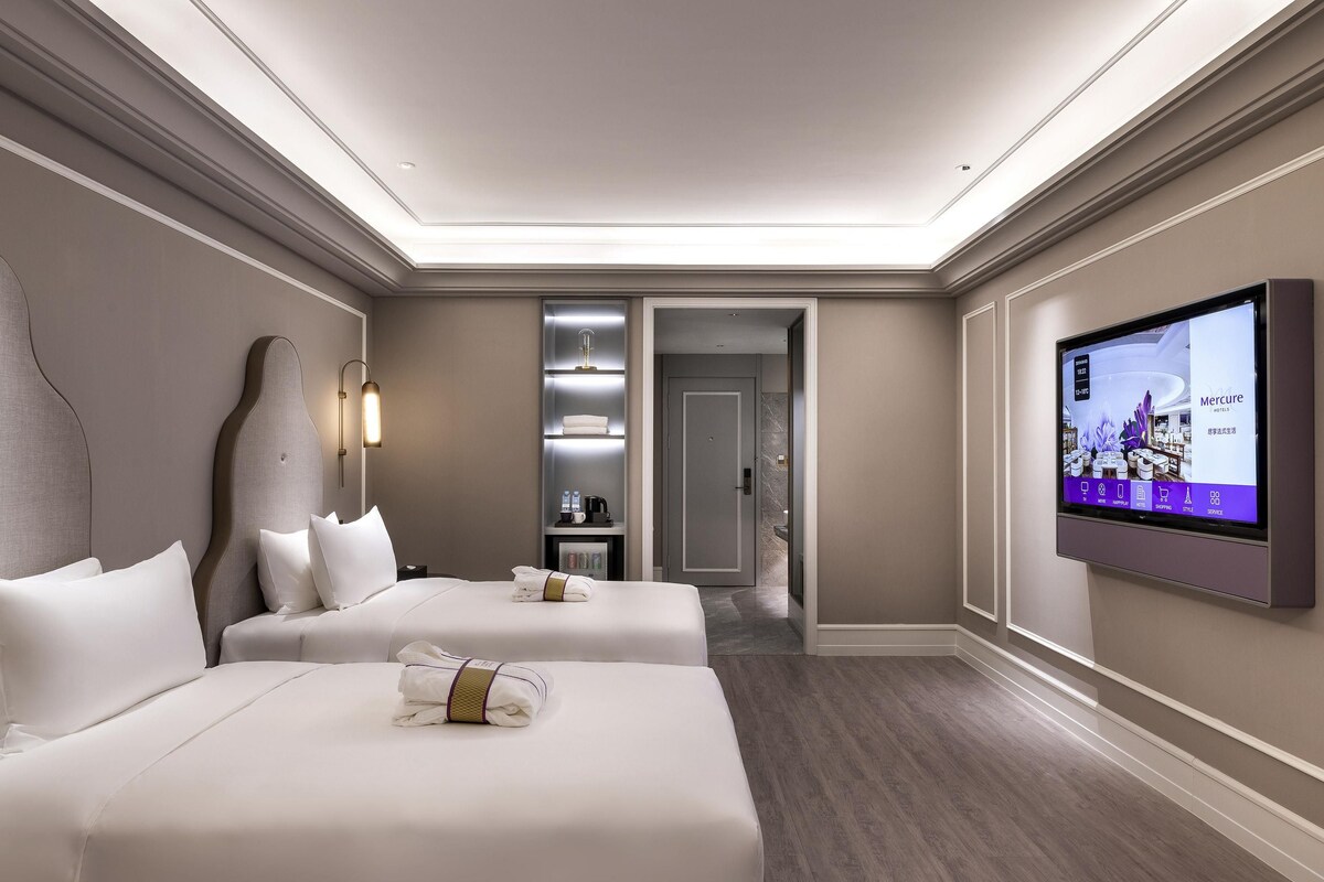 A hotel room with two beds, a television, a minibar, and a bathroom visible through an open door.