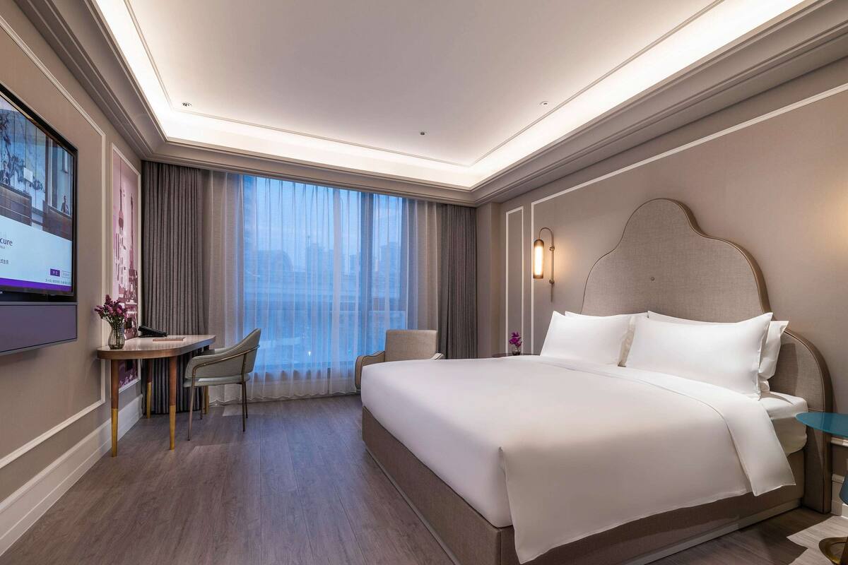 Een hotelkamer met een groot bed, een bureau met stoel, een televisie aan de muur en een raam met gordijnen.