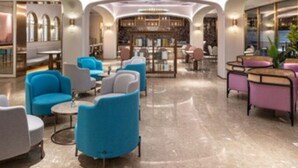 Een moderne lobby met een marmeren vloer, blauwe en grijze fauteuils, en een centraal zitgedeelte met een ronde lamp.