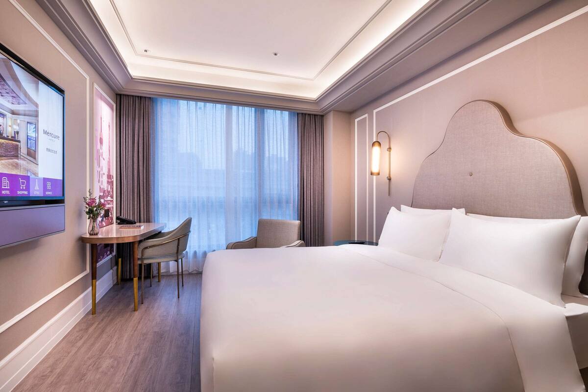 Une chambre d’hôtel avec un grand lit, un bureau avec une chaise, une télévision et une fenêtre avec des rideaux.