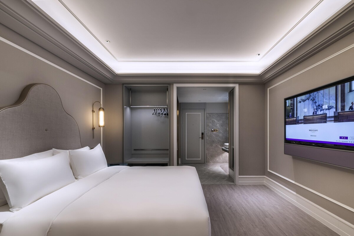 A hotel room with a large bed, a television, a wardrobe, and a bathroom visible through an open door.
