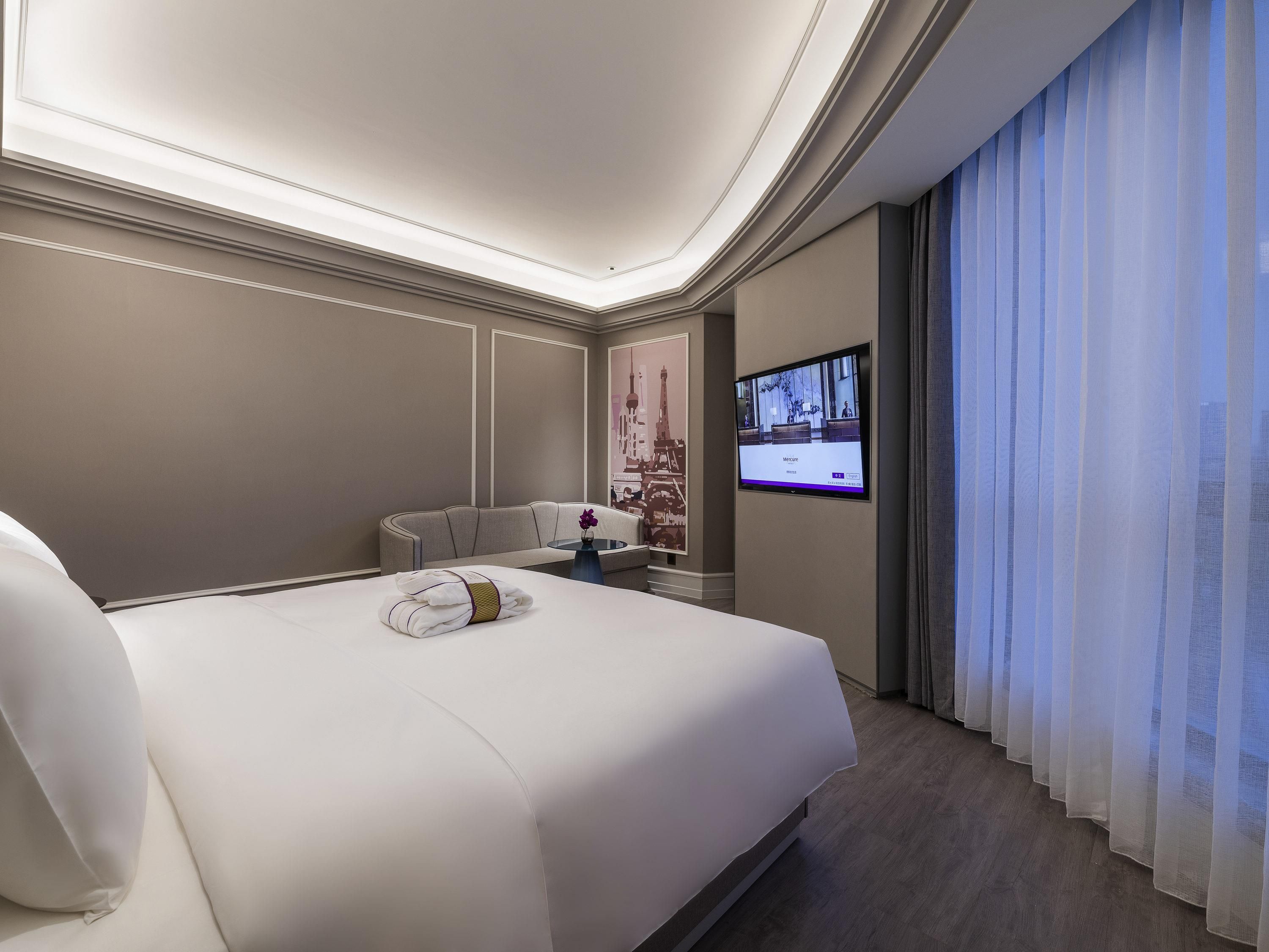 A hotel room with a large bed, a sofa, a TV mounted on the wall, and a window with sheer curtains.