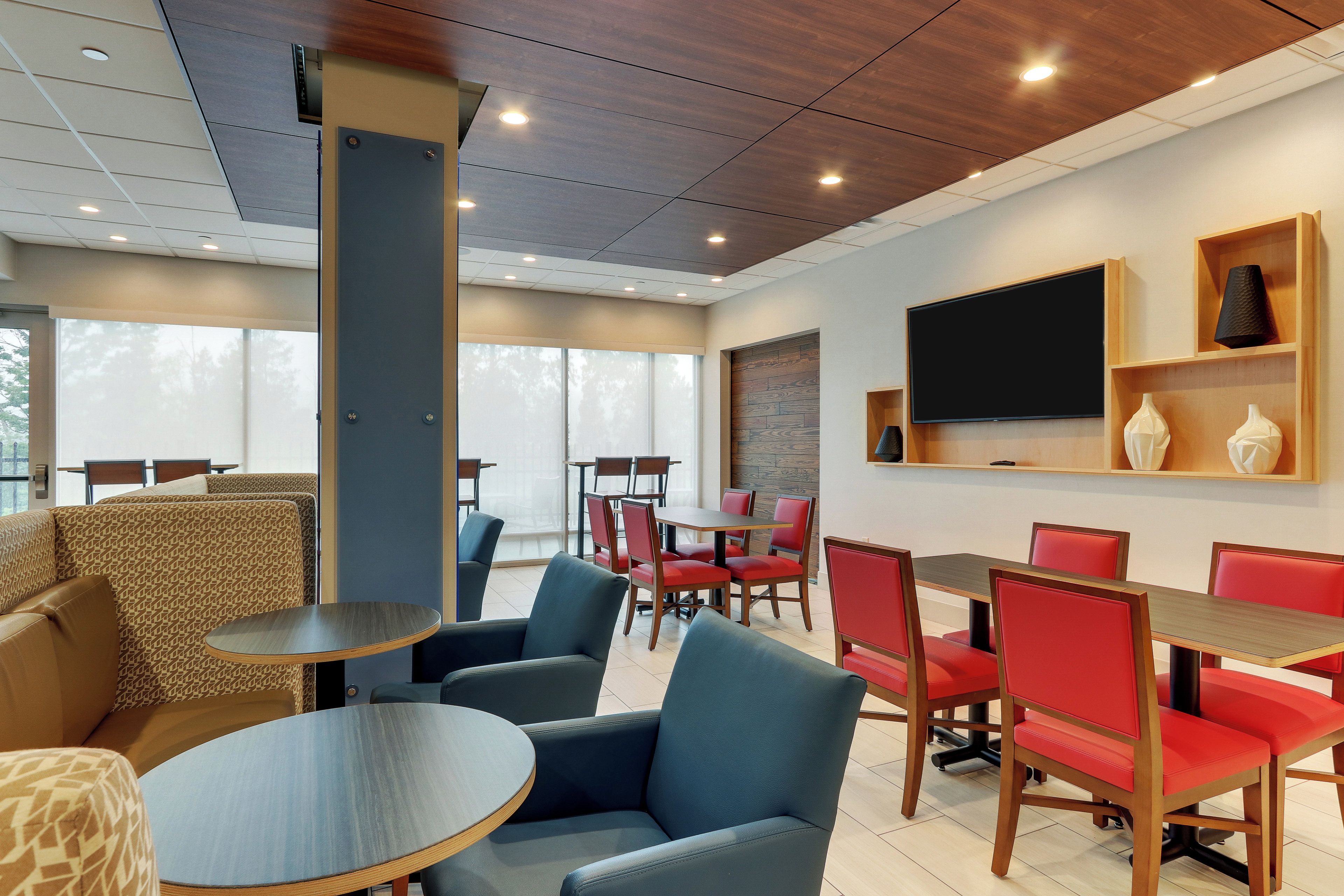 A hotel room with a large window, a flat-screen TV mounted on the wall, and a seating area with tables and chairs.