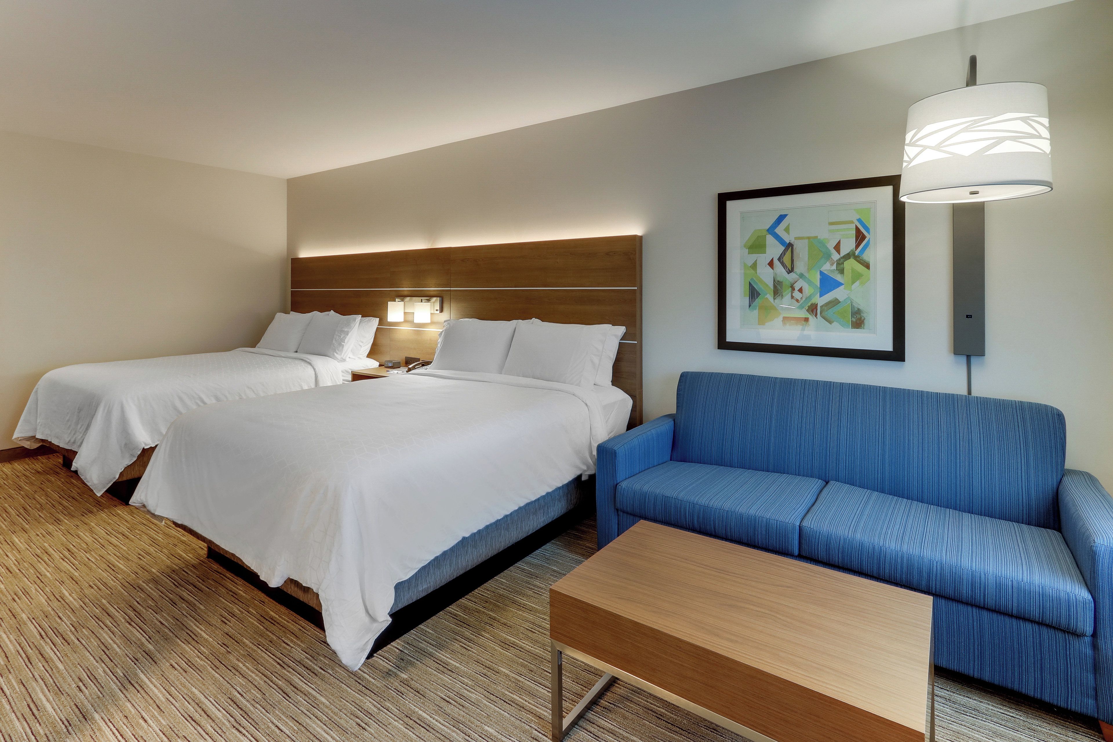 Una habitación de hotel con dos camas, un sofá azul, una mesa de centro de madera y un cuadro abstracto enmarcado en la pared.