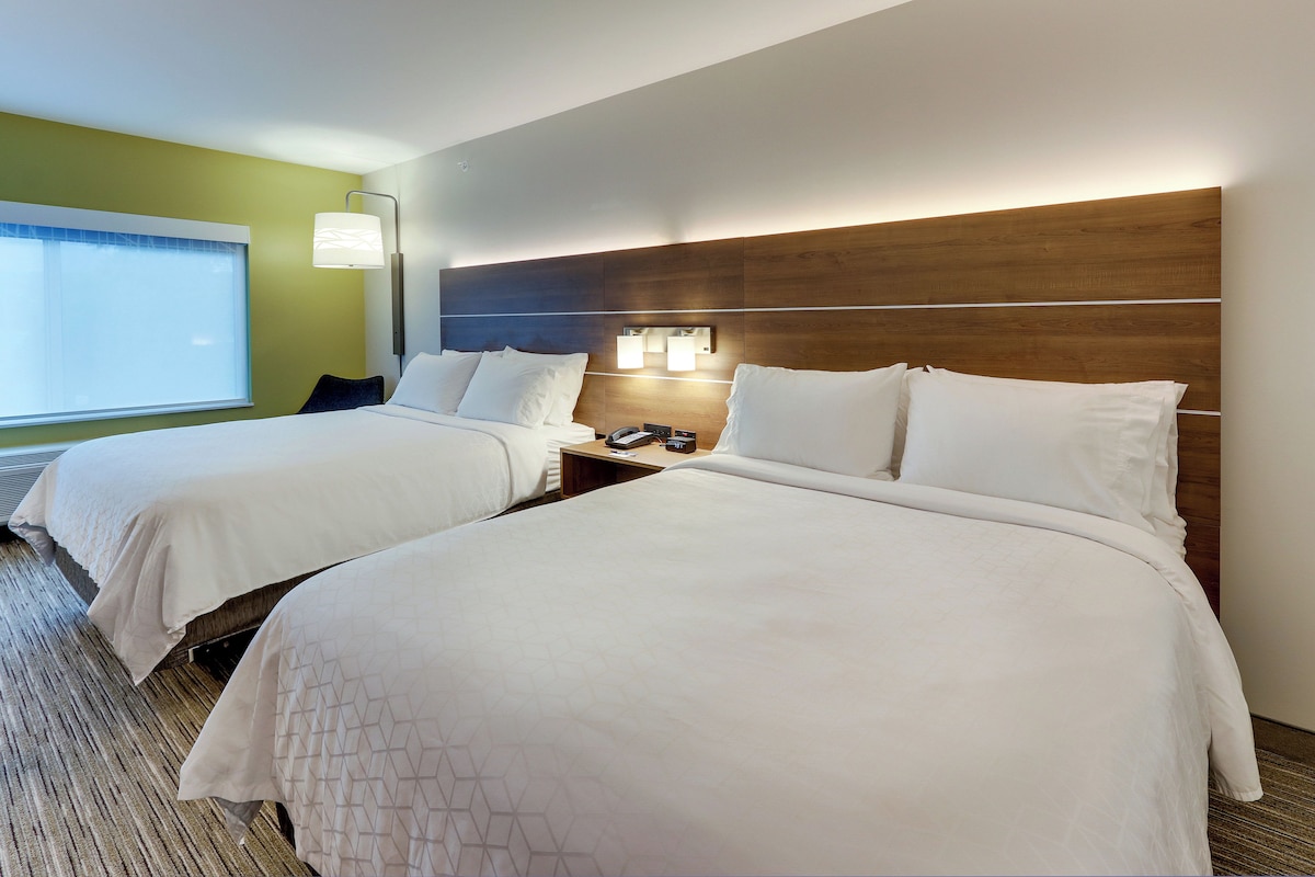 Une chambre d’hôtel avec deux lits, une table de chevet avec une lampe et une fenêtre avec des stores.