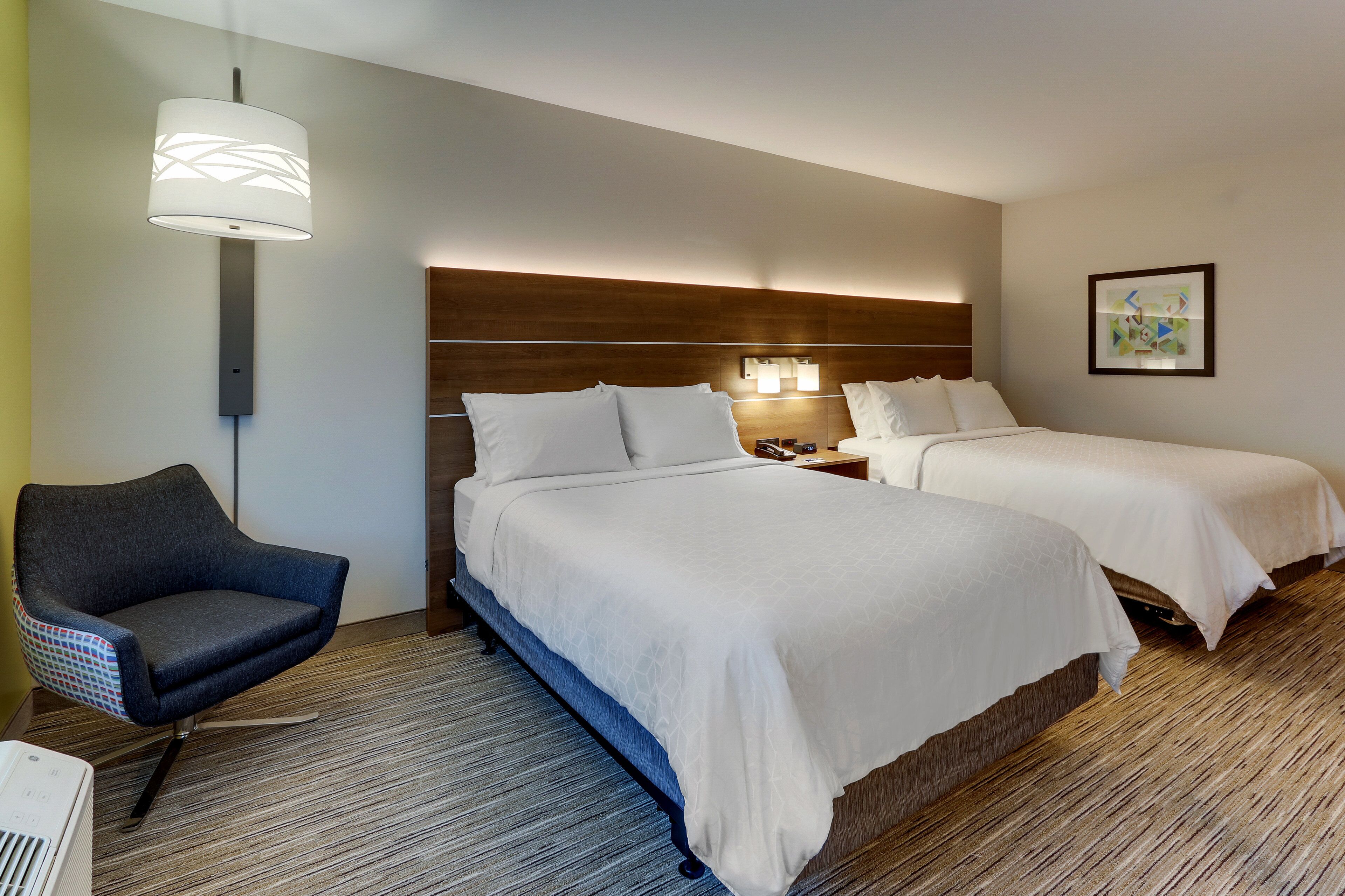 A hotel room with two beds, a chair, a nightstand, and a wall-mounted lamp.