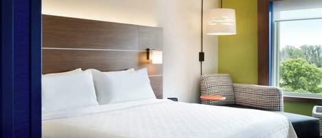 Habitación de hotel con una cama grande, una mesita de noche, una lámpara, una silla y una ventana con vistas a árboles.
