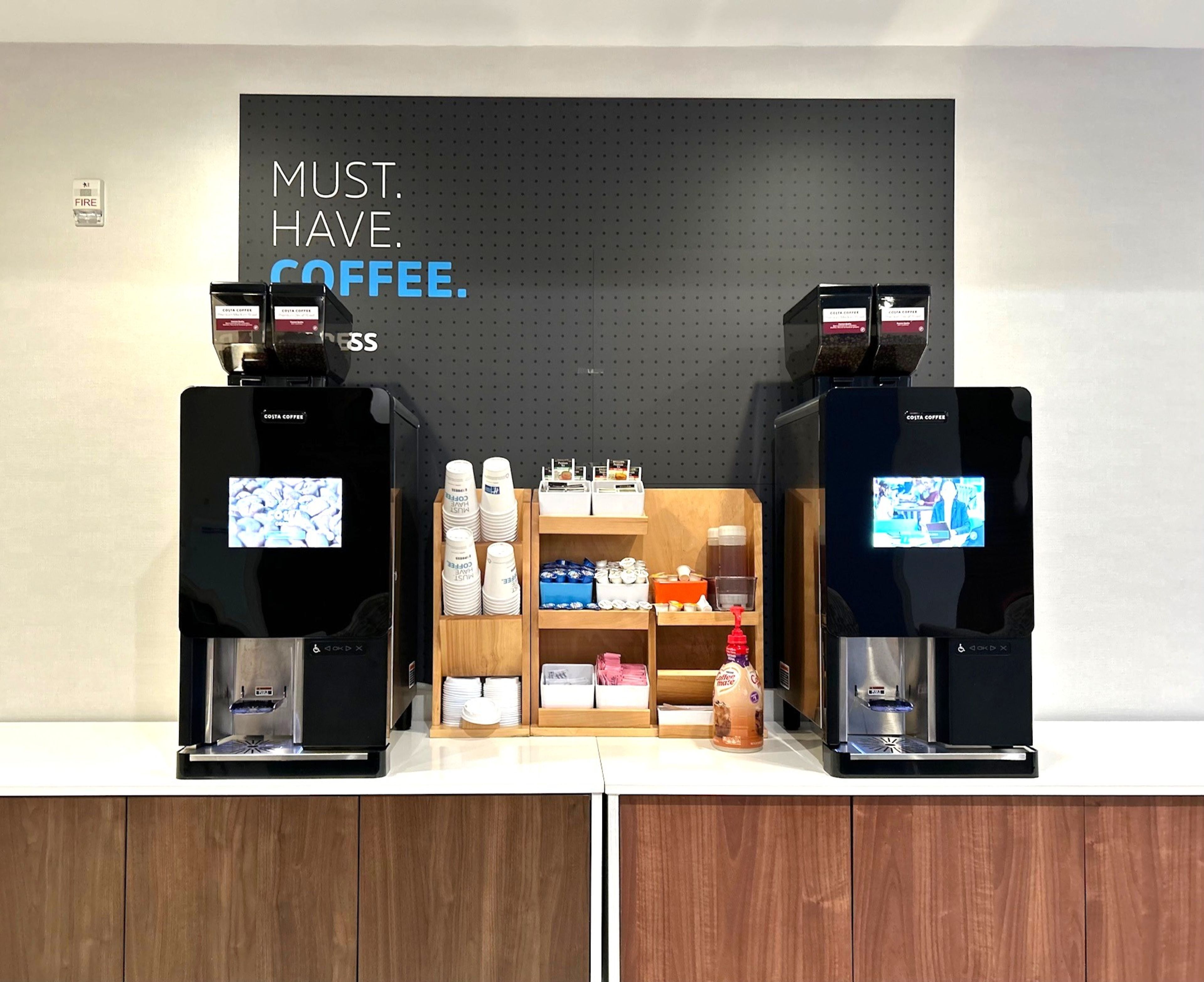 Un espace dédié au café, équipé de deux machines à café, d’un détecteur d’incendie et d’une étagère contenant des tasses et des sachets de café.