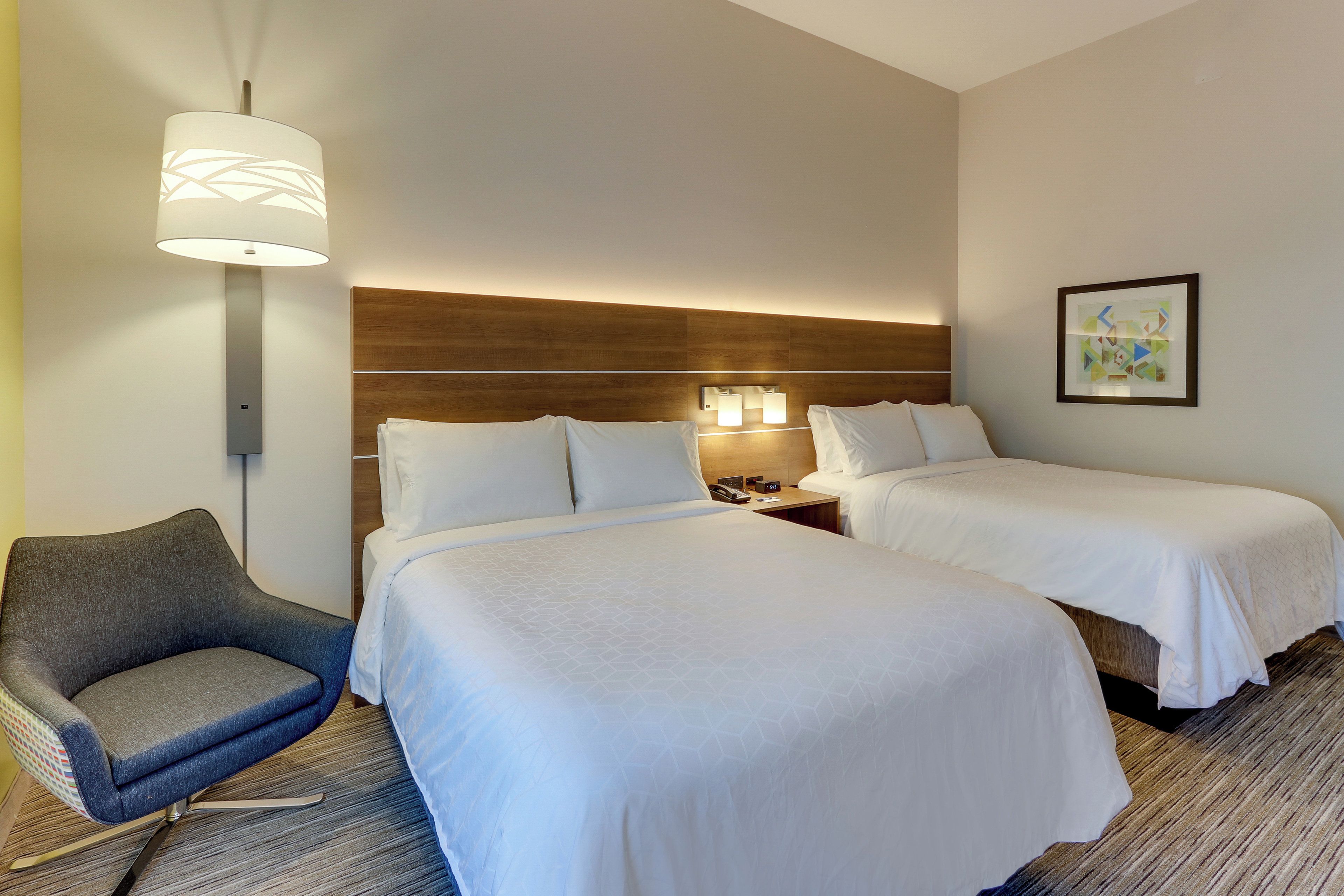Quarto de hotel com duas camas, uma cadeira, mesa de cabeceira com luminária e quadro na parede.