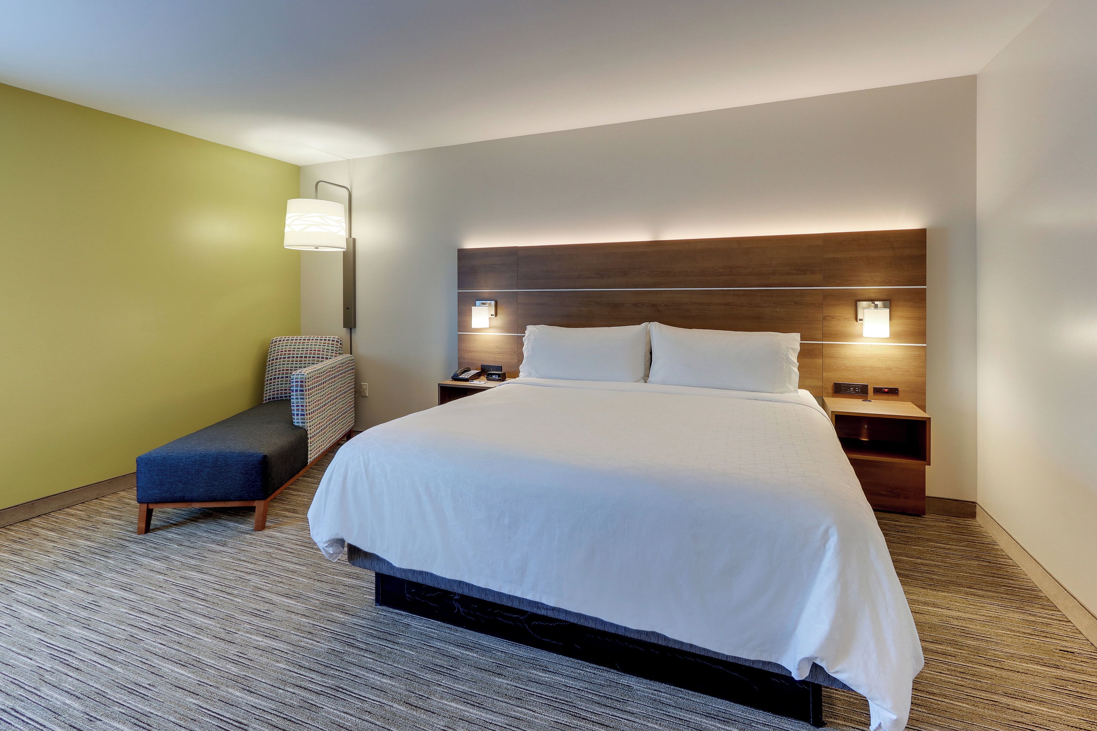 Una habitación de hotel con una cama grande, mesitas de noche, un banco y una lámpara de pared.