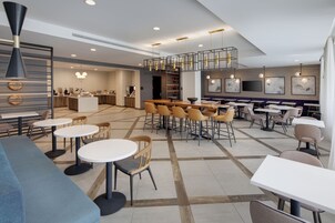 Un restaurante moderno con mesas y sillas redondas, una zona de cocina y un área de estar con techo alto e iluminación colgante.