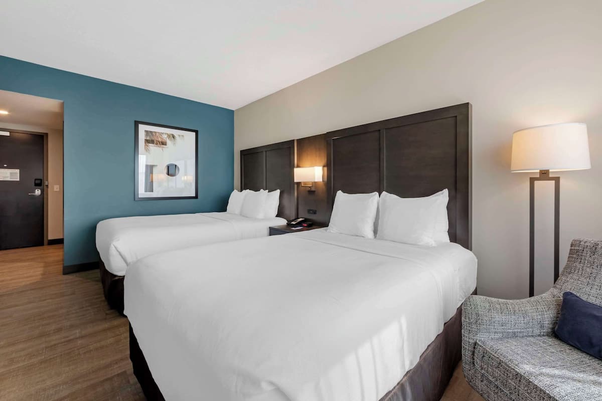 A hotel room with two beds, a blue accent wall, a framed picture, a nightstand with a lamp, and a chair.