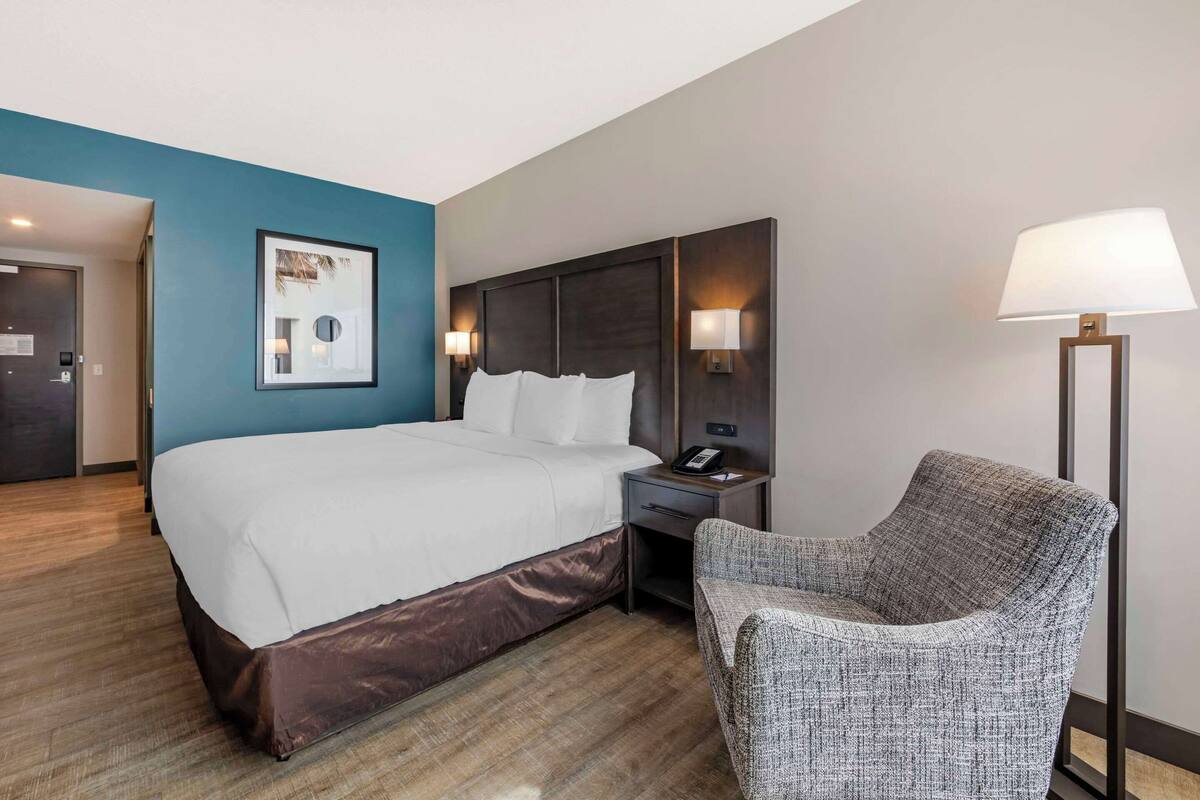 A hotel room with a large bed, a chair, a desk, a lamp, and a wall-mounted artwork.