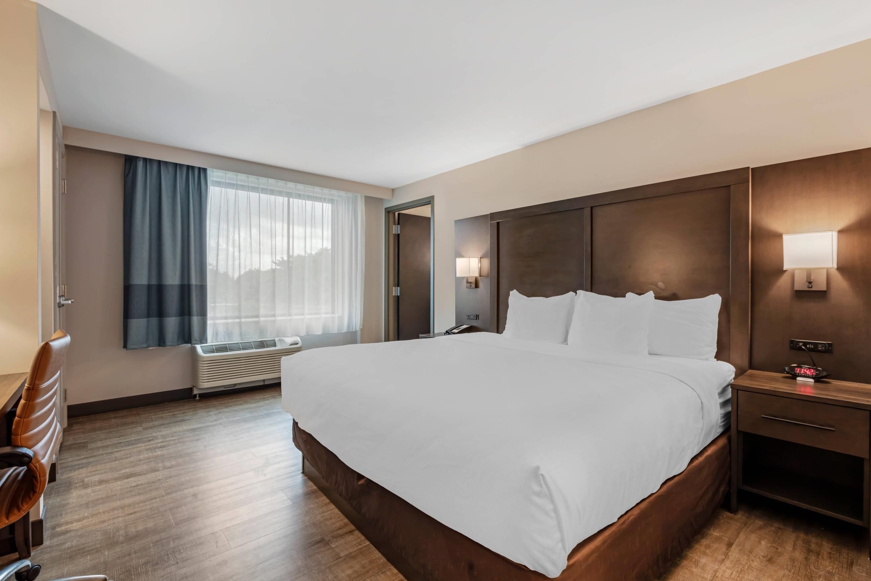 A hotel room with a large bed, bedside tables, a desk, a chair, a window with curtains, and a wall-mounted lamp.