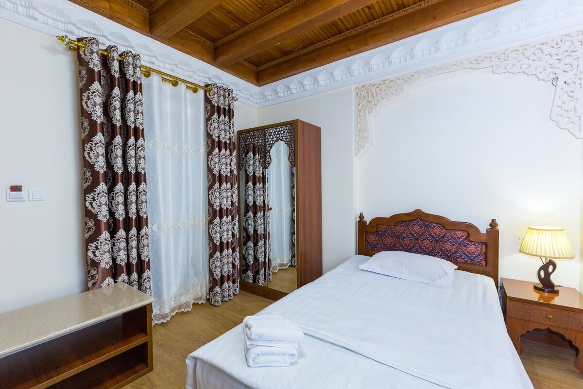 Un dormitorio con una cama de madera, sábanas blancas, una mesita de noche con lámpara y un banco.