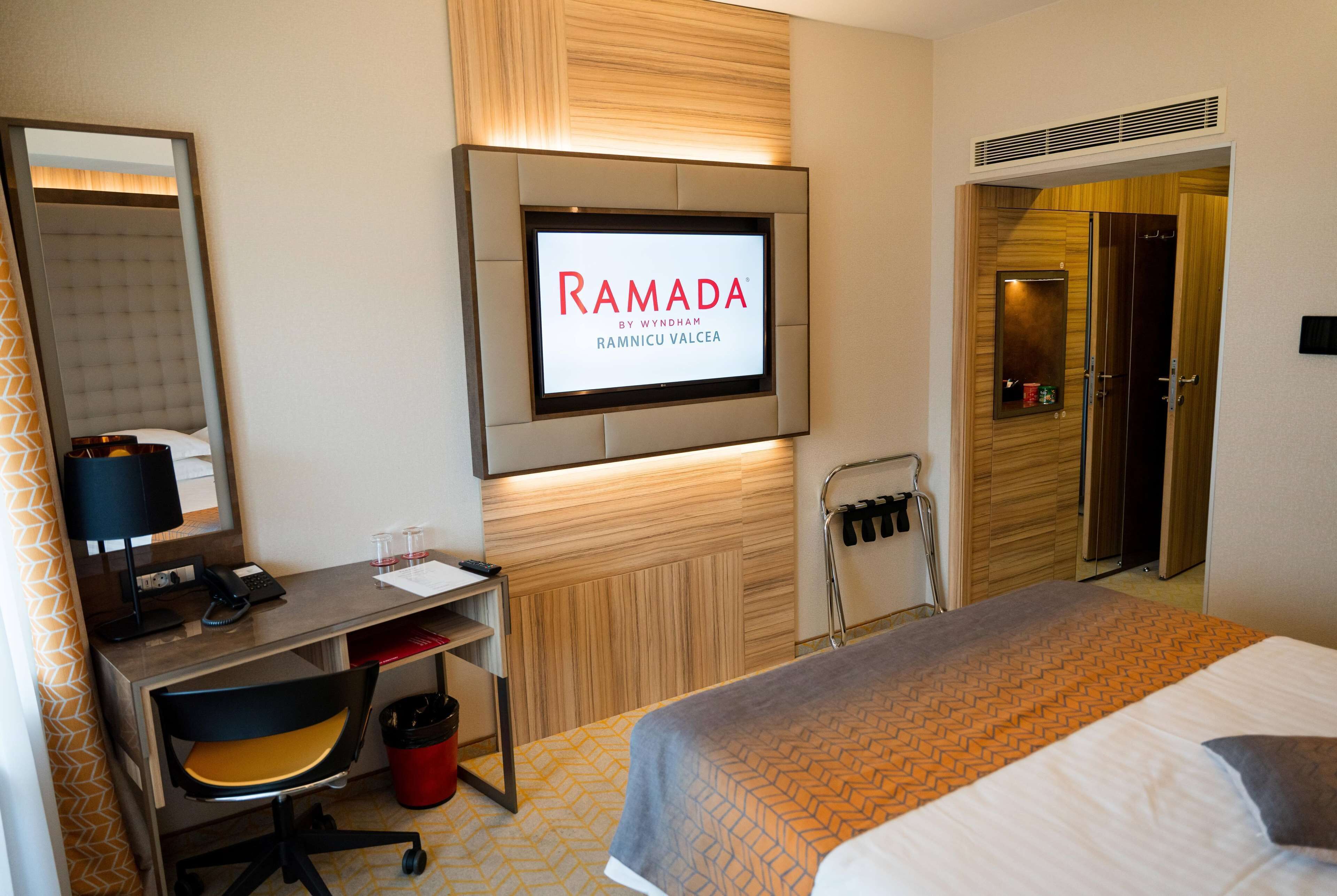 A hotel room with a bed, a desk with a computer, a TV mounted on the wall, and a bathroom door.