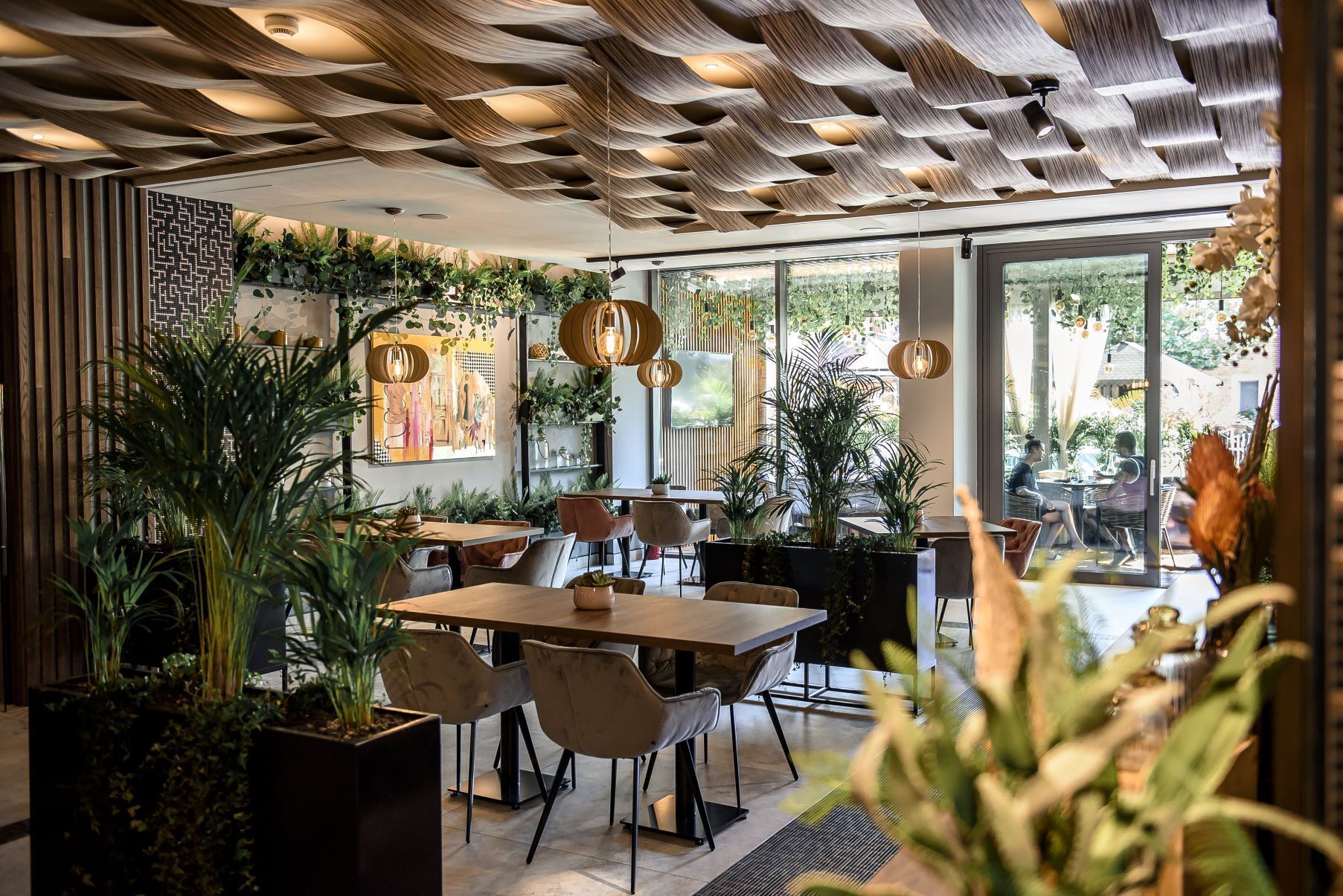 A modern dining area with wooden tables and chairs, surrounded by potted plants and a patterned ceiling.