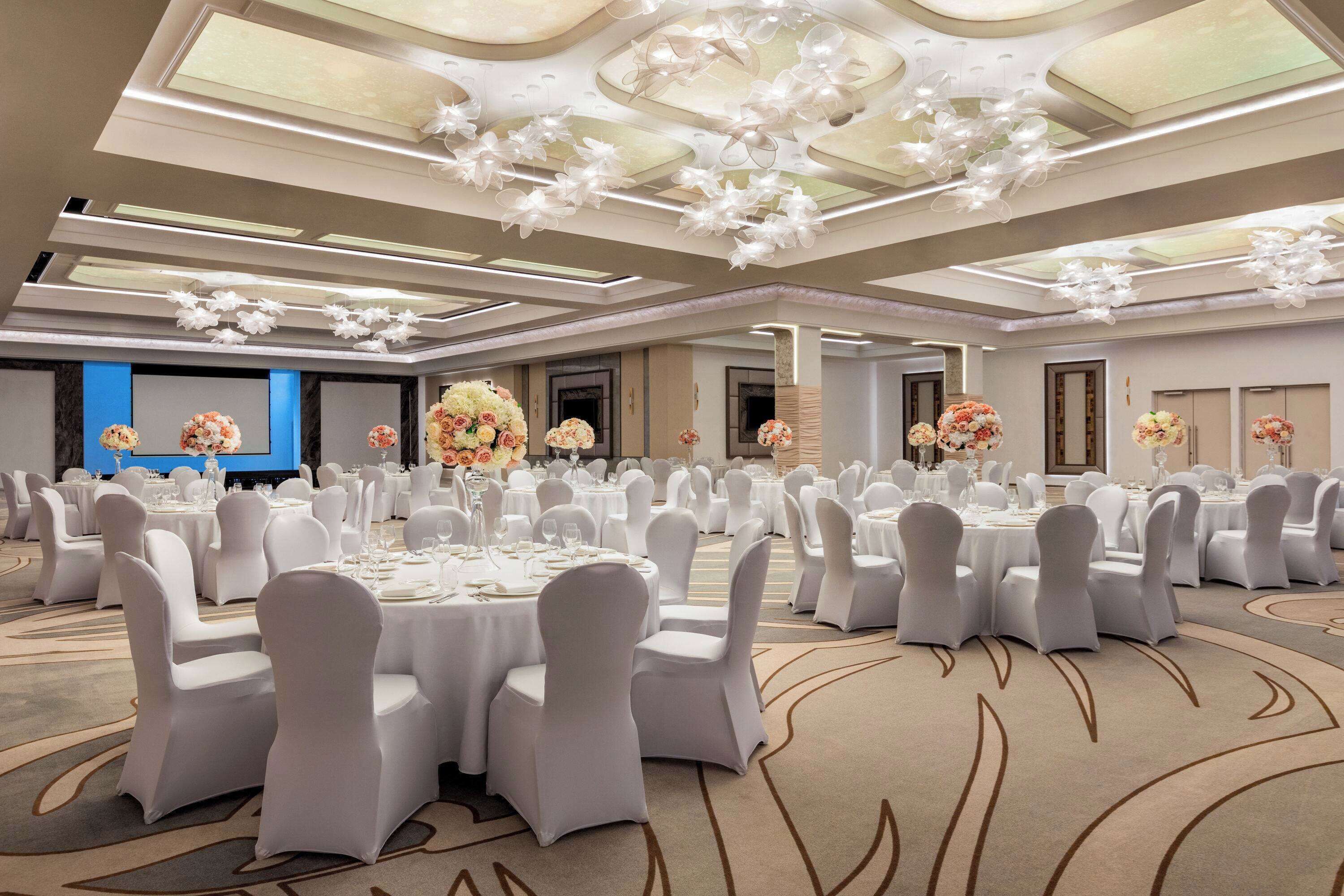 Een ruime balzaal met ronde tafels, witte stoelen en bloemdecoraties.