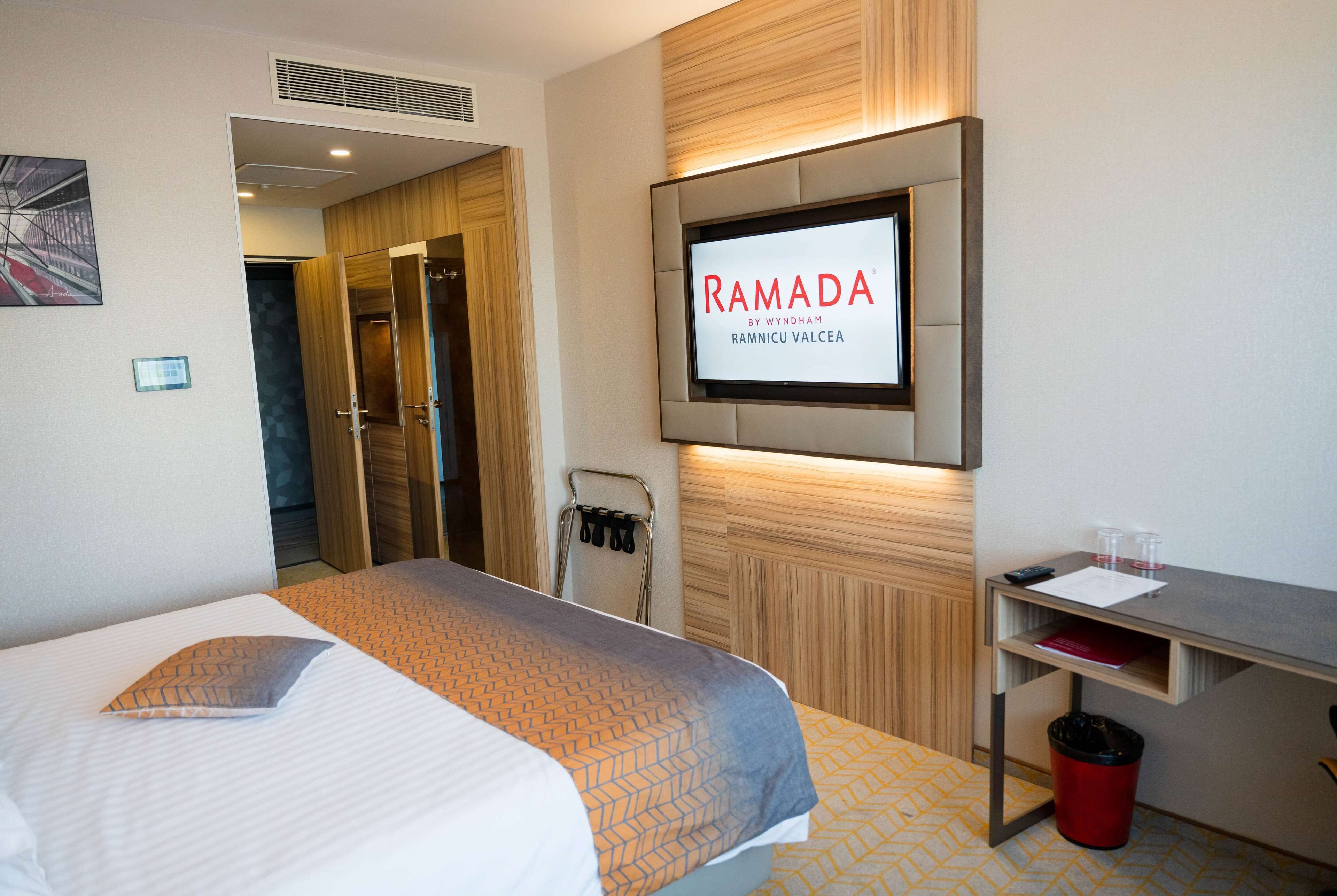 Habitación de hotel con cama, televisión montada en la pared, escritorio con cubo de basura y puerta que da al baño.