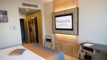 Habitación de hotel con cama, televisión montada en la pared, escritorio con cubo de basura y puerta que da al baño.