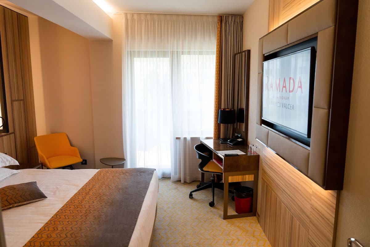 Une chambre d’hôtel avec un lit, un bureau, une chaise, une télévision et une fenêtre avec des rideaux.