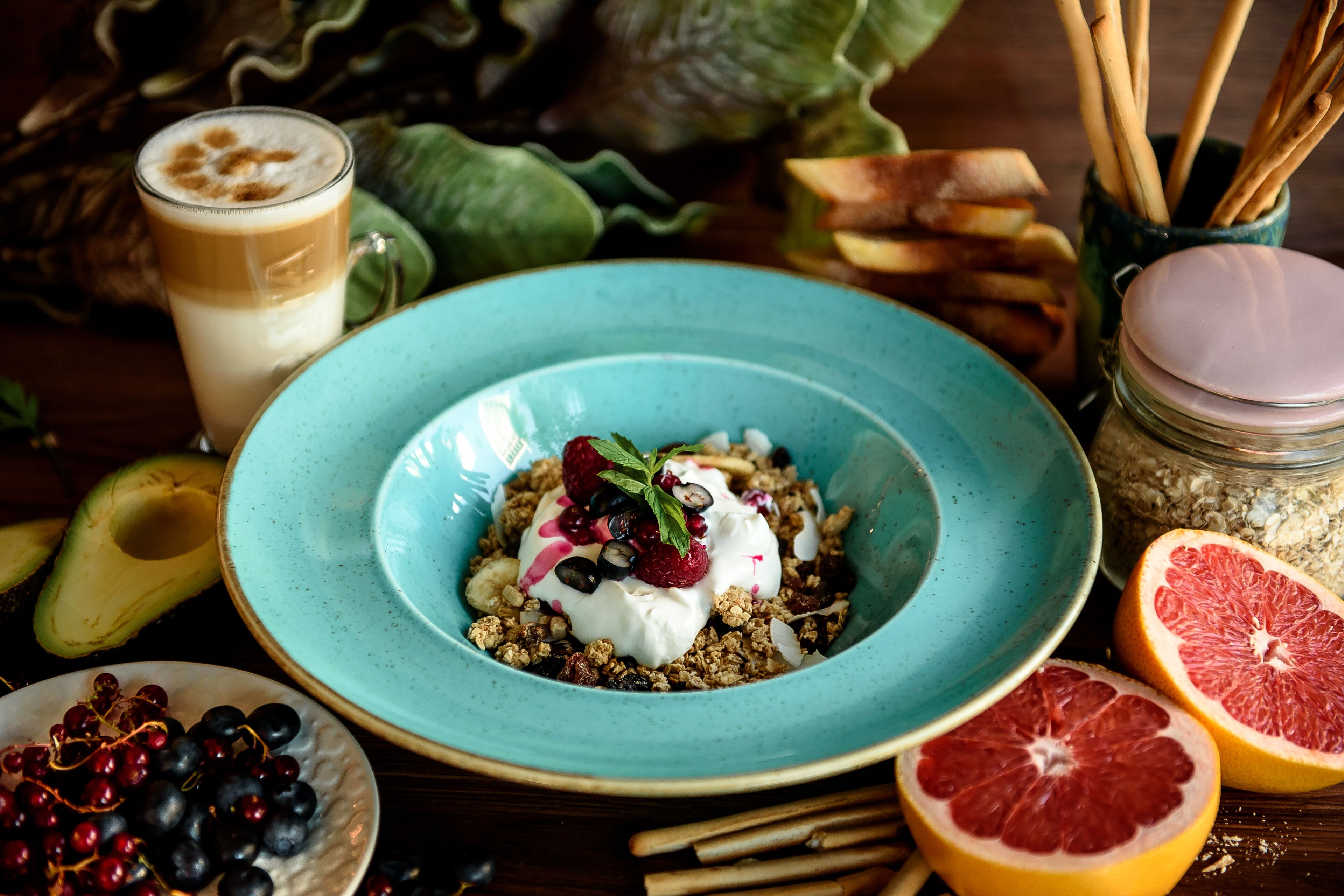 A breakfast setting with a bowl of granola topped with yogurt and berries, a glass of coffee, and fresh fruits like avocado and grapefruit.