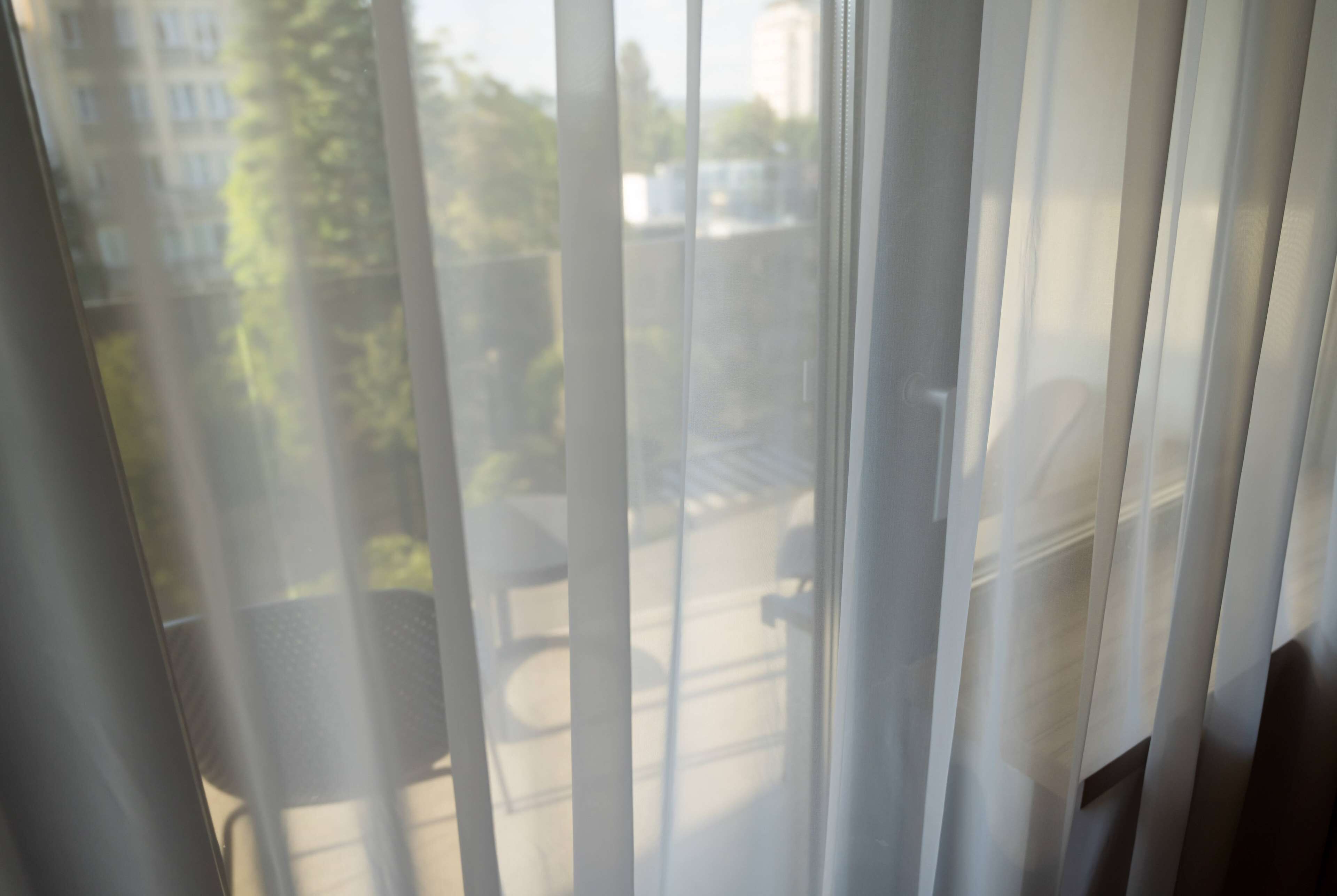 Blick durch transparente Vorhänge, zeigt eine Straßenszene mit Gebäuden und Grünflächen.