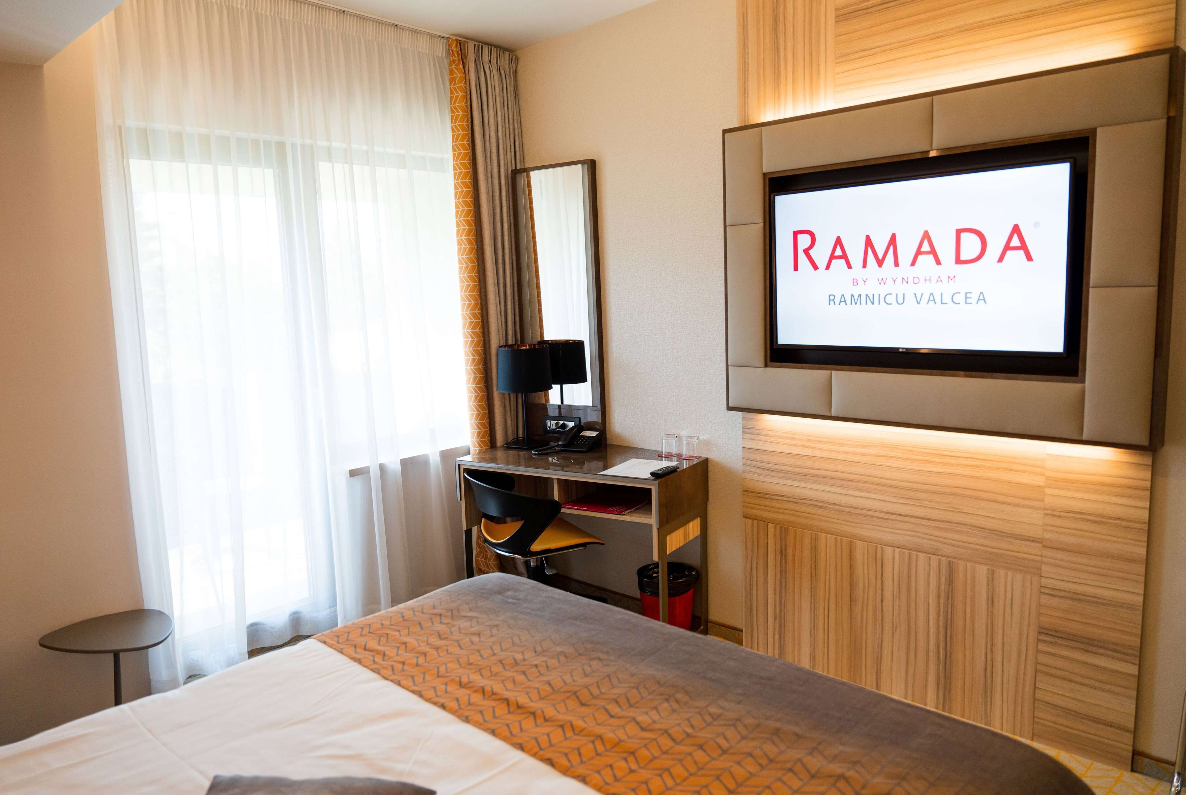 A hotel room with a bed, a desk with a computer, a TV mounted on the wall, and a window with curtains.