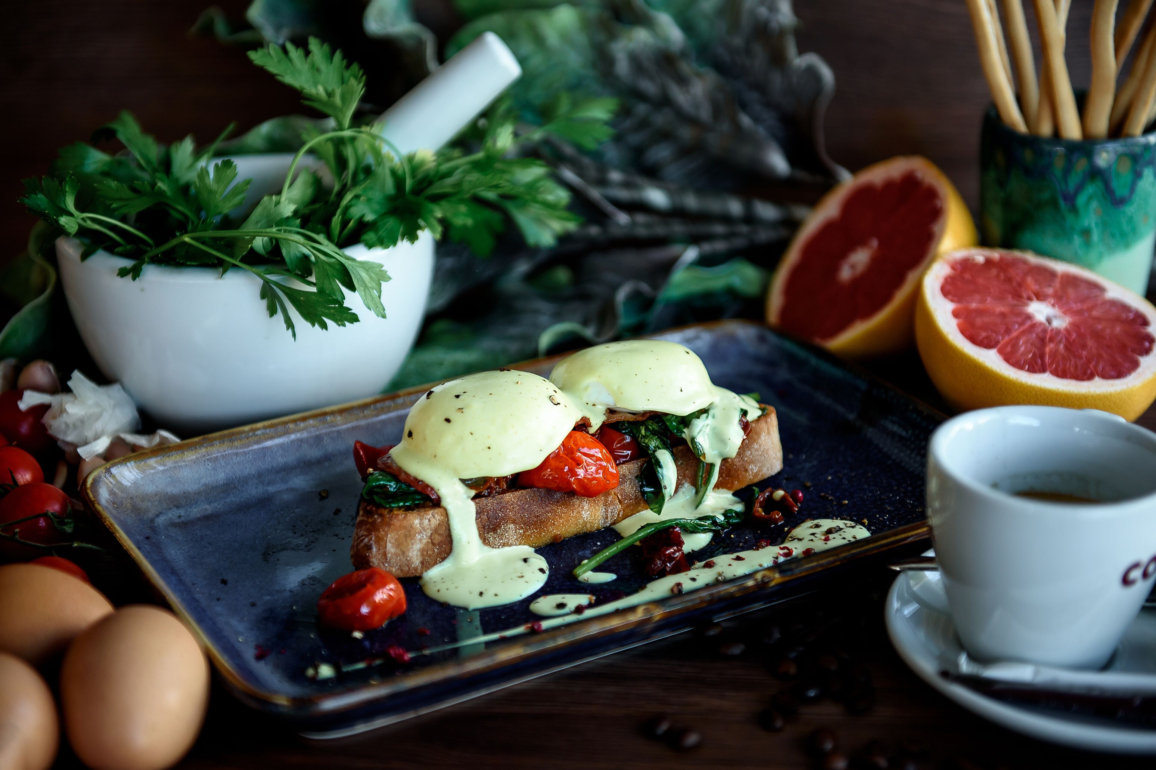 Un plato de desayuno con huevos, tomates y verduras de hoja, un bol de perejil, media toronja y una taza de café.