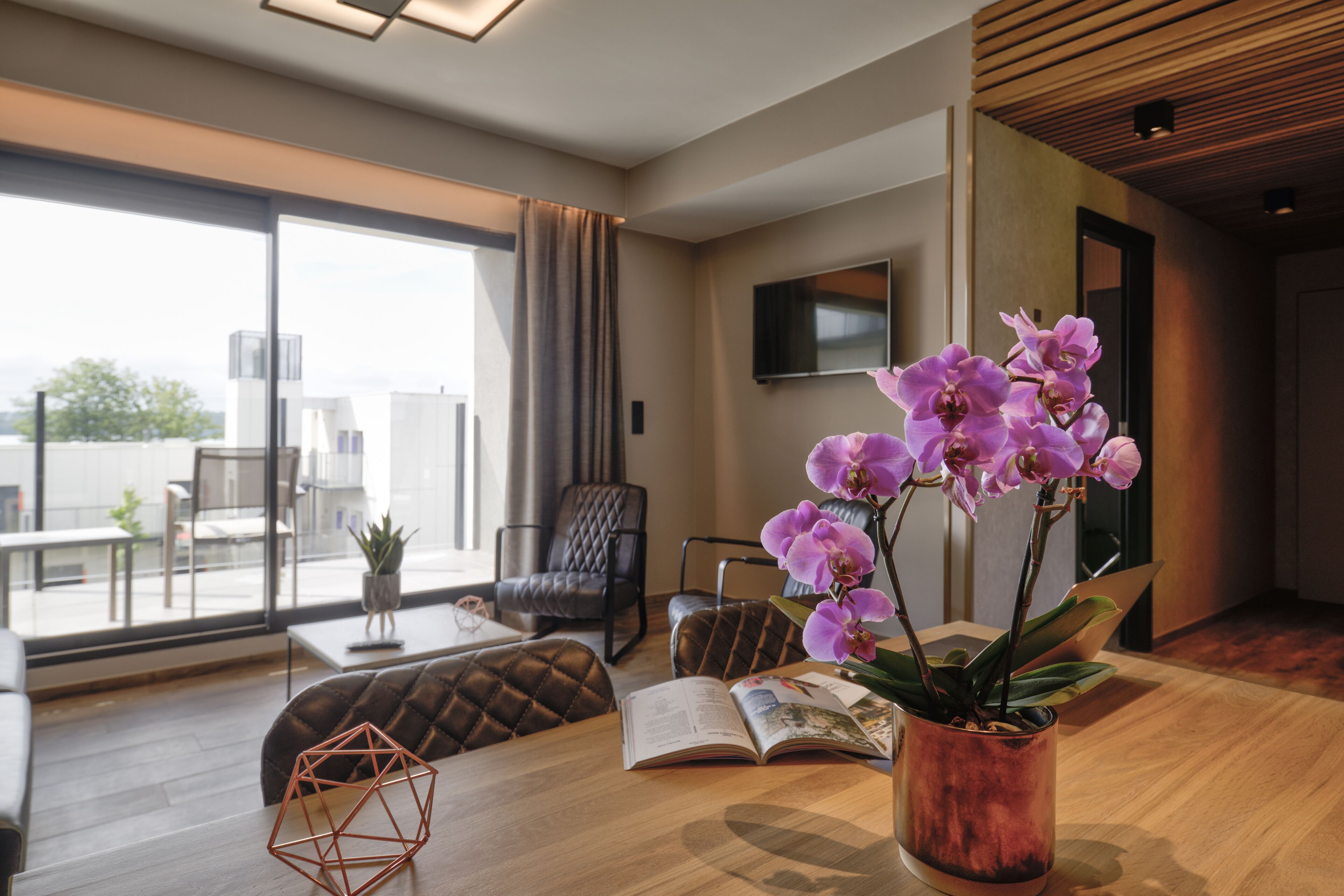 A modern living room with a large window, a wooden coffee table, a leather sofa, and a potted orchid plant.
