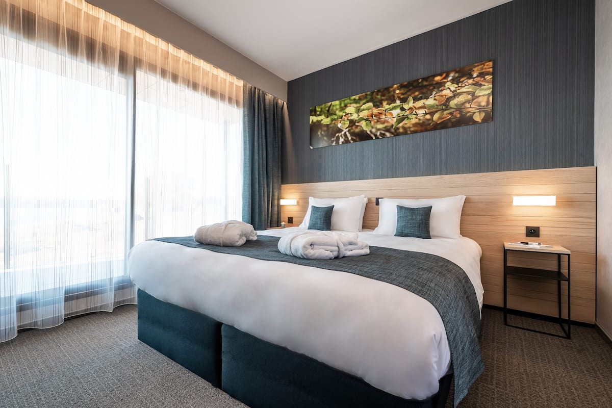 A modern hotel room with a large bed, a headboard with integrated lighting, a side table, and a large window with sheer curtains.