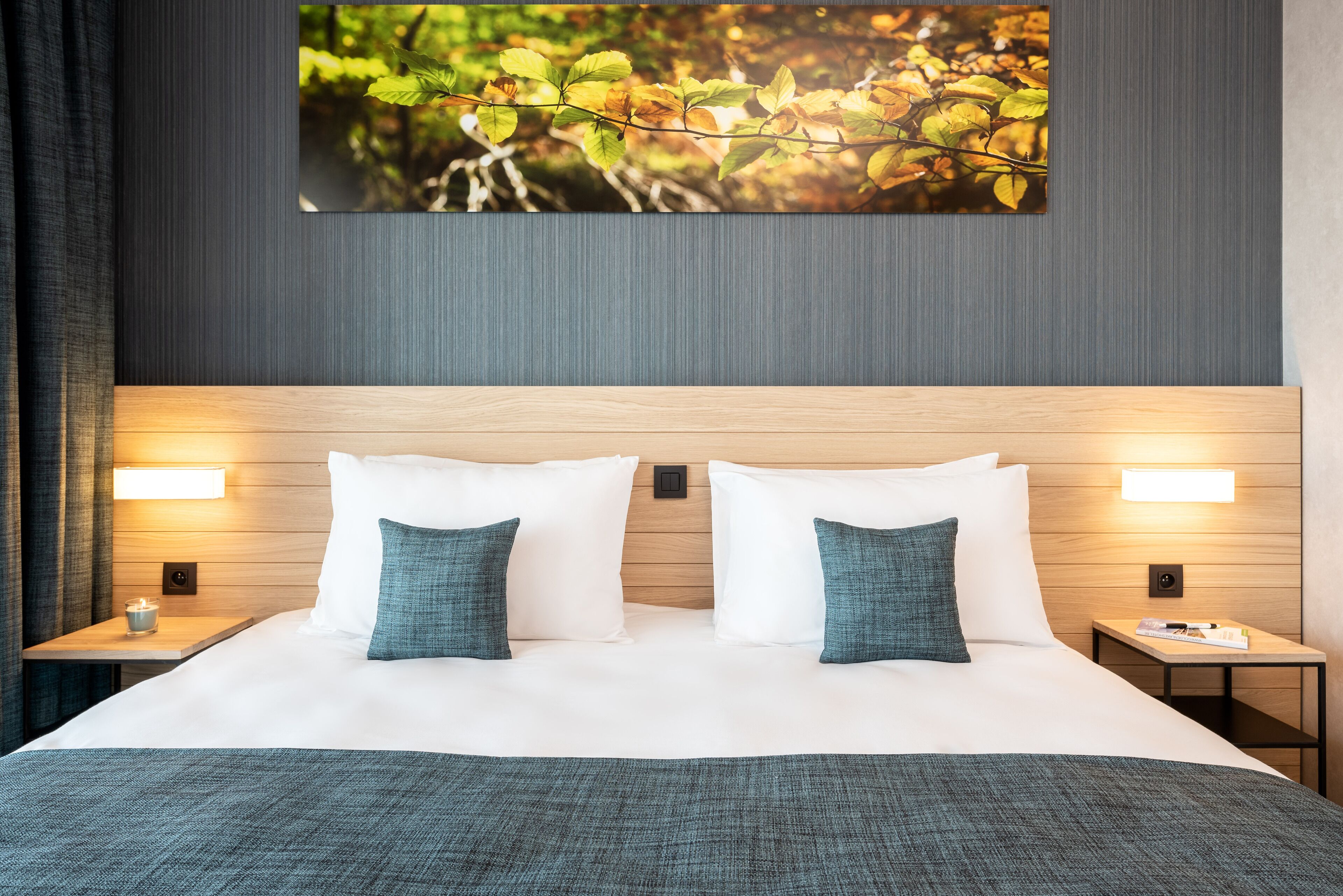 Una cama impecablemente vestida con almohadas blancas y azul verdosas, un cabecero de madera y dos mesitas de noche con lámparas.