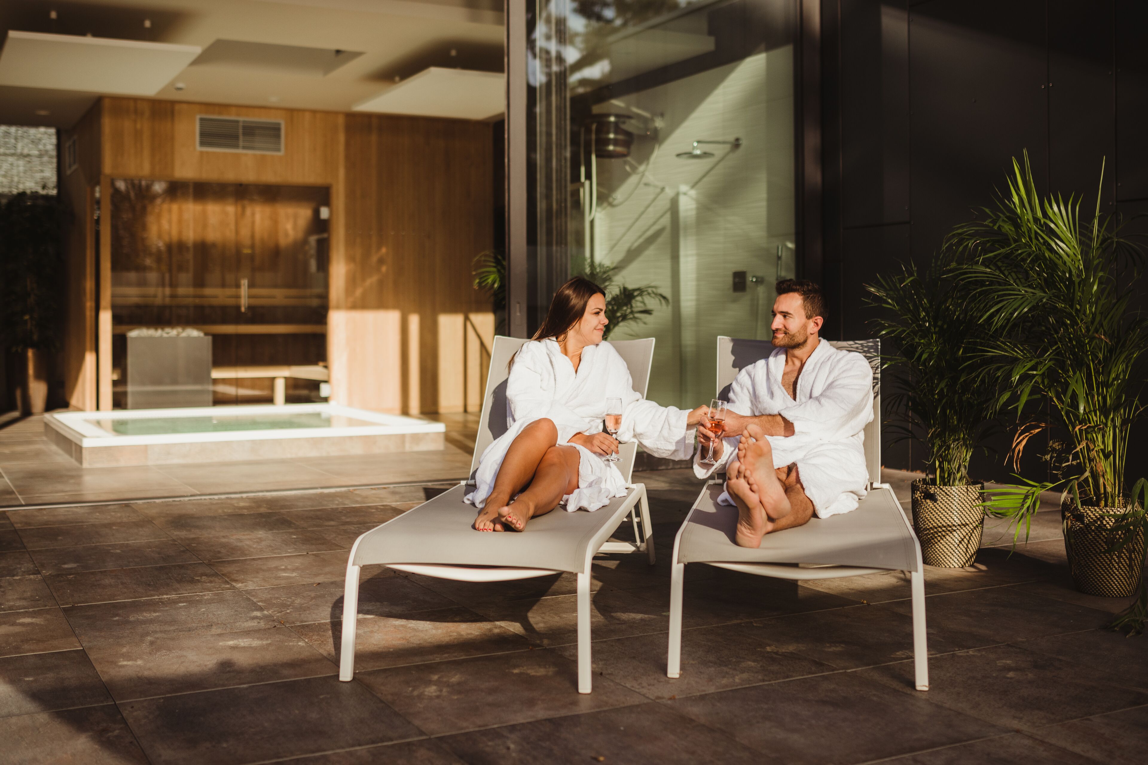 Two people sitting on lounge chairs in a modern outdoor area with a glass wall and a pool in the background.