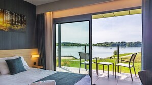 A hotel room with a large bed, a view of a lake, and a balcony with outdoor seating. - Golden Lakes Hotel (Froidchapelle)