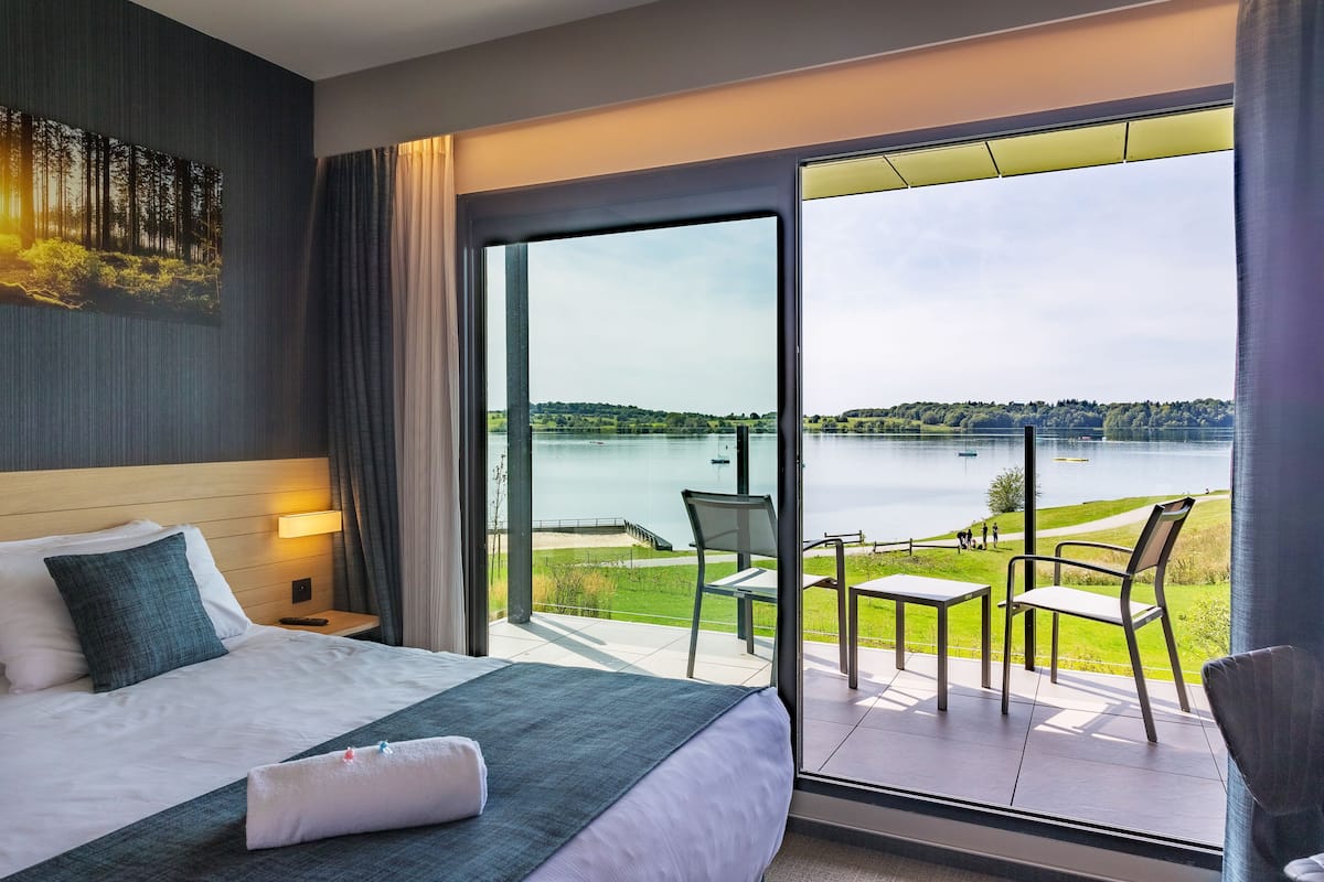 A hotel room with a large bed, a view of a lake, and a balcony with outdoor seating.