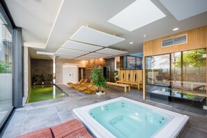 A modern indoor pool area with a hot tub, lounge chairs, and a glass-enclosed patio. - Golden Lakes Hotel (Froidchapelle)