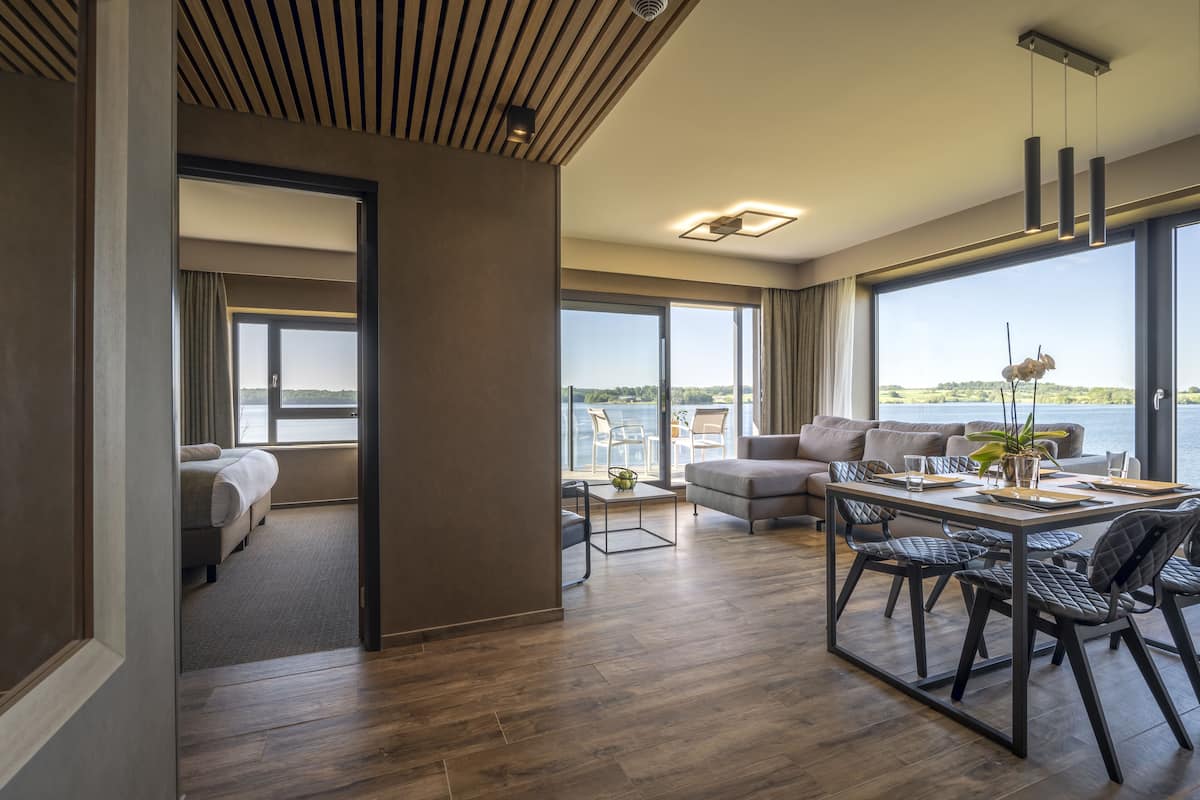 A modern hotel room with a large balcony, a dining area with a table and chairs, and a cozy sitting area with a sofa.