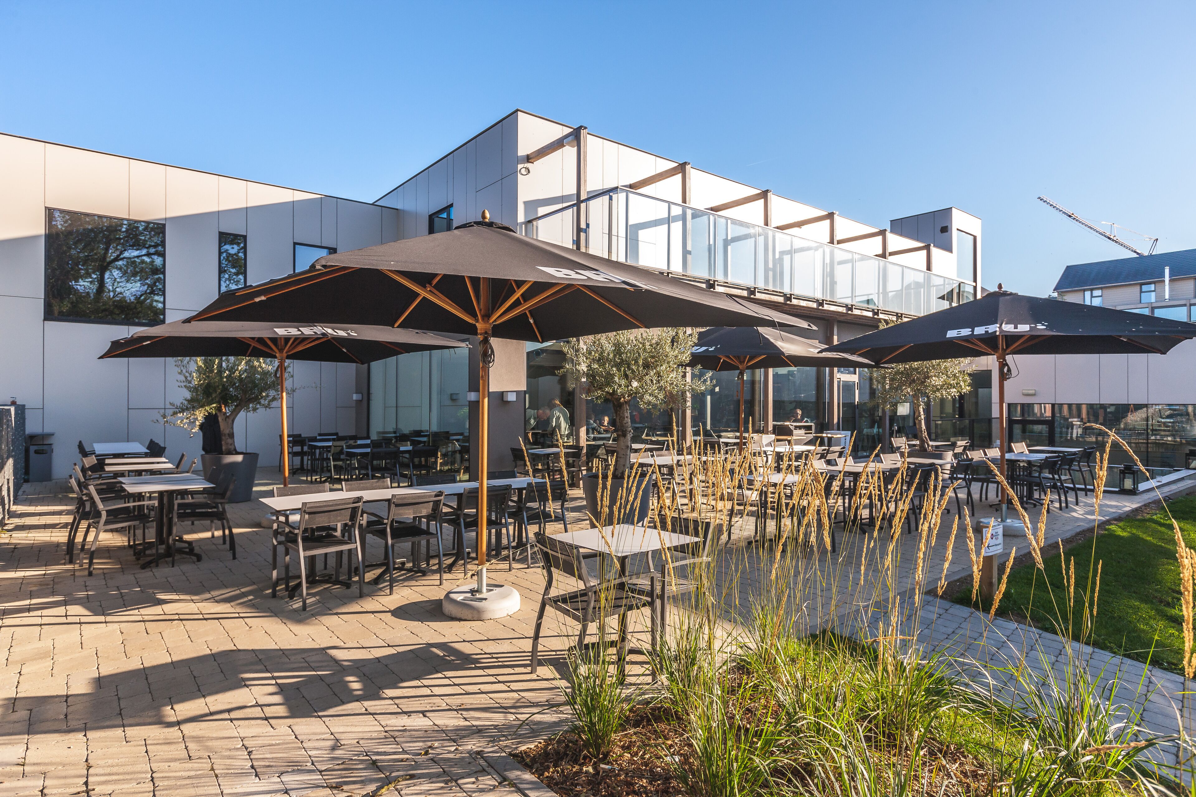 Un área de comedor al aire libre con mesas y sillas, sombrillas y un edificio moderno al fondo.