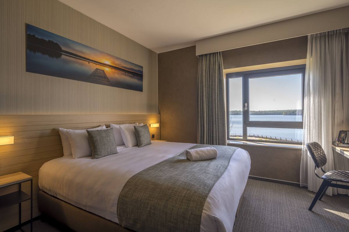 A hotel room with a large bed, a view of the water, a desk, and a chair.
