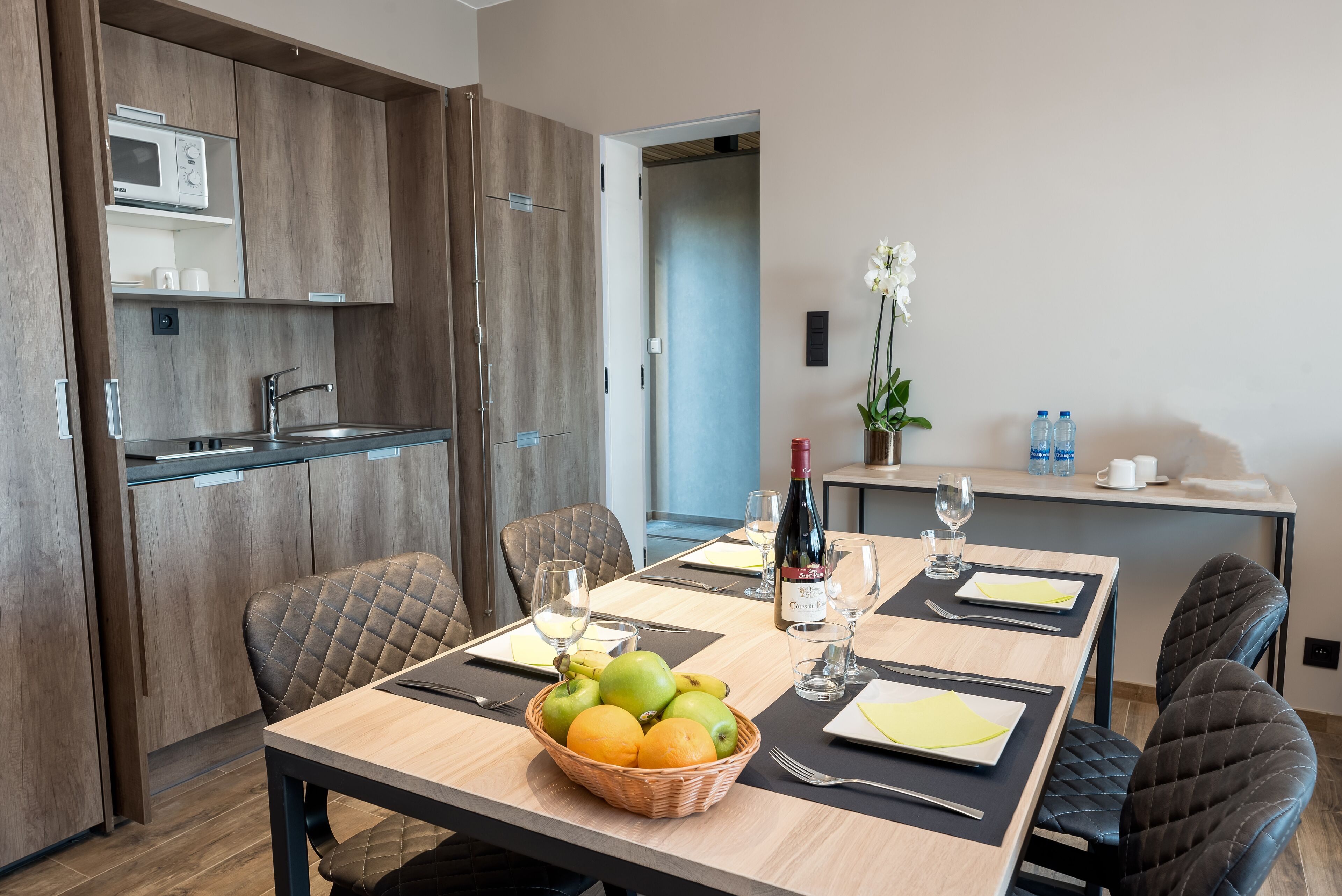 Un coin repas moderne avec une table en bois dressée pour quatre personnes, un panier de fruits et une bouteille de vin.