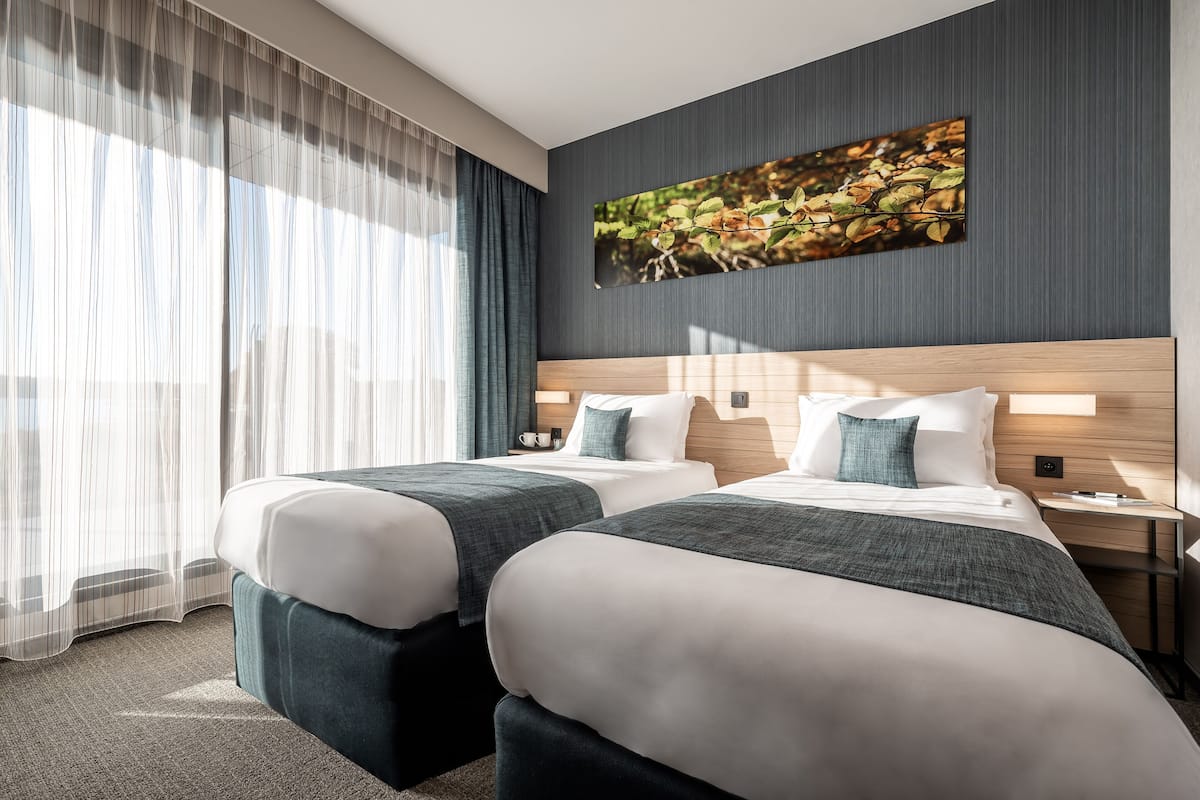 A hotel room with two beds, a large window with sheer curtains, a wall-mounted painting, and a nightstand with a lamp.