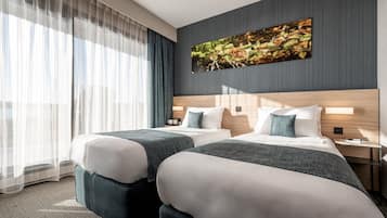 A hotel room with two beds, a large window with sheer curtains, a wall-mounted painting, and a nightstand with a lamp.