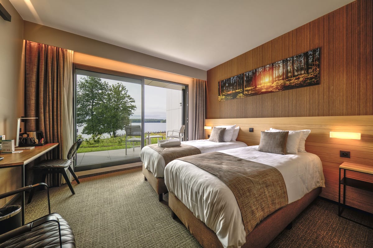 A hotel room with two beds, a desk, and a chair. There is a large window with a view of trees and a body of water.