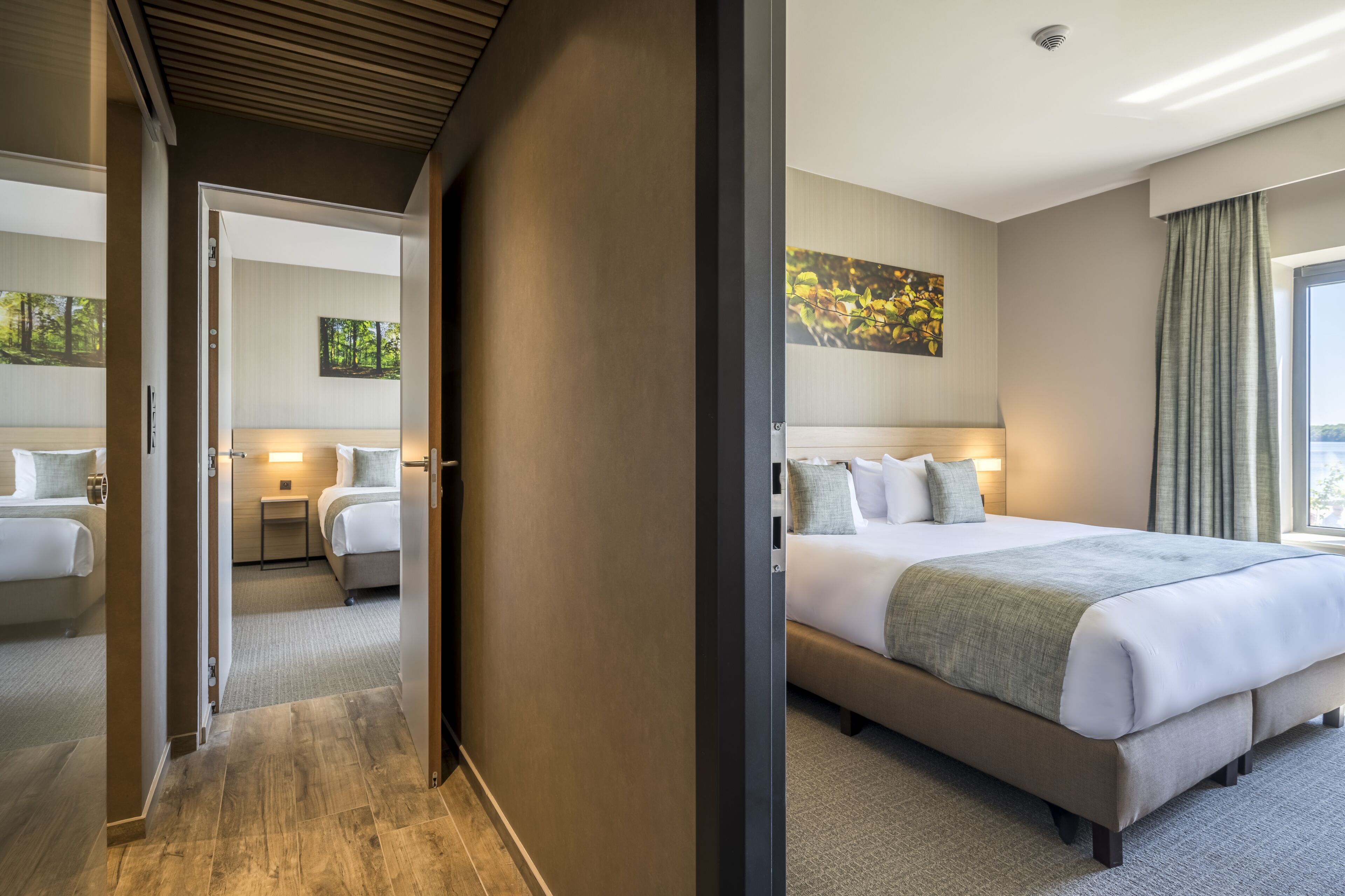 Quarto de hotel com uma cama grande, mesa de cabeceira, criado-mudo e um quadro na parede.