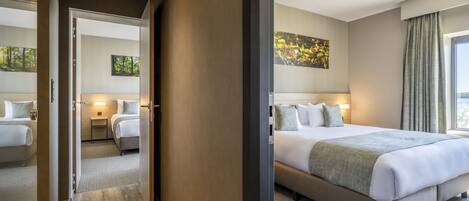 Quarto de hotel com uma cama grande, mesa de cabeceira, criado-mudo e um quadro na parede.