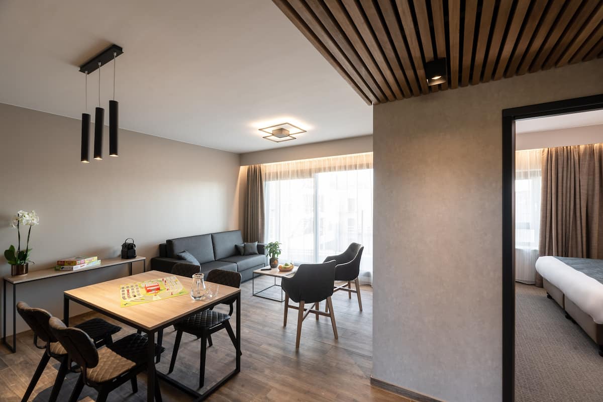 A modern hotel room with a wooden dining table, black chairs, a sofa, and a bed.