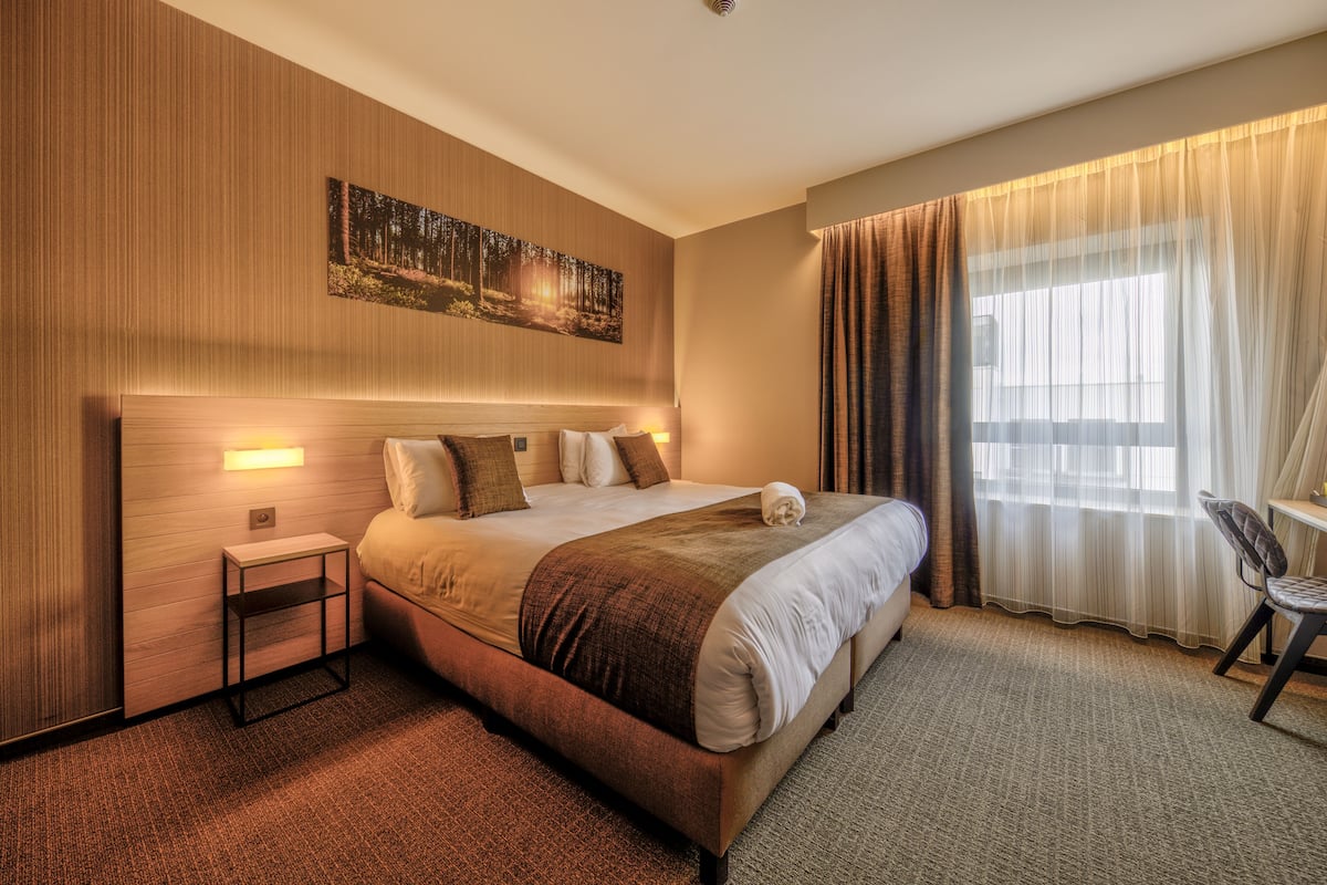 A hotel room with a large bed, a bedside table, a chair, and a window with curtains.