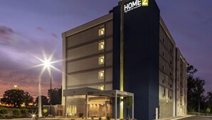 Un hotel de varios pisos con un llamativo letrero 'HOME 2', un acceso cubierto y un área de estacionamiento.